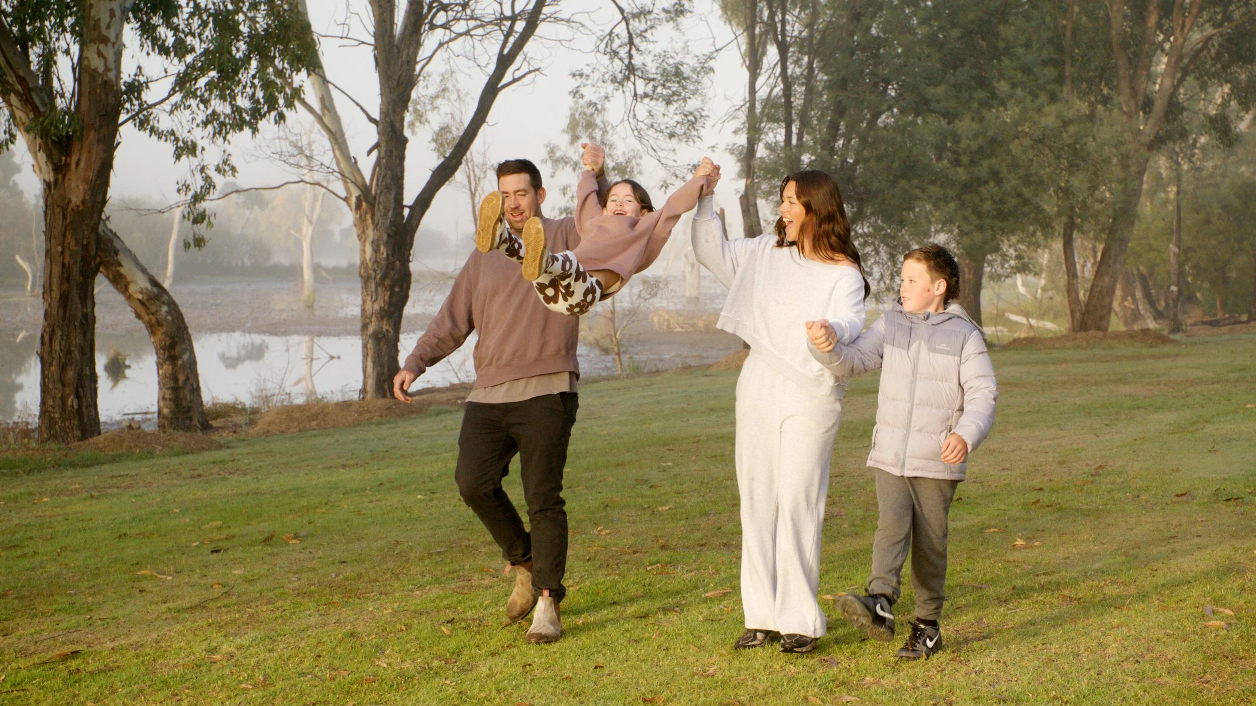 Family enjoying a walk in a park with trees and a lake in the background, with the parents lifting their daughter and all of them smiling.