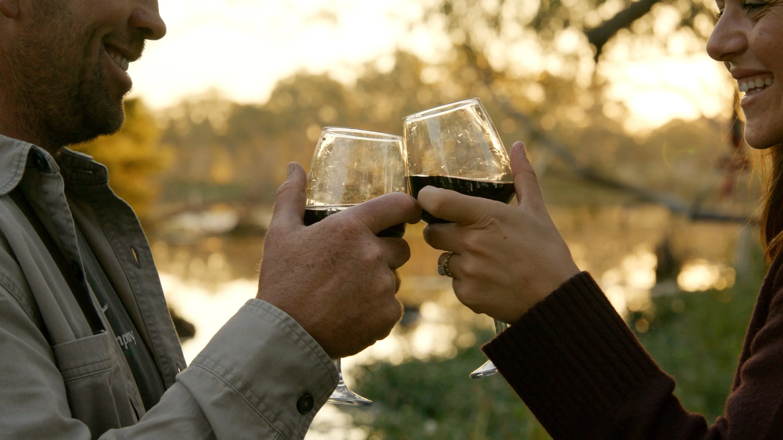 Two people clink glasses of red wine outdoors during sunset, smiling and enjoying each other's company.