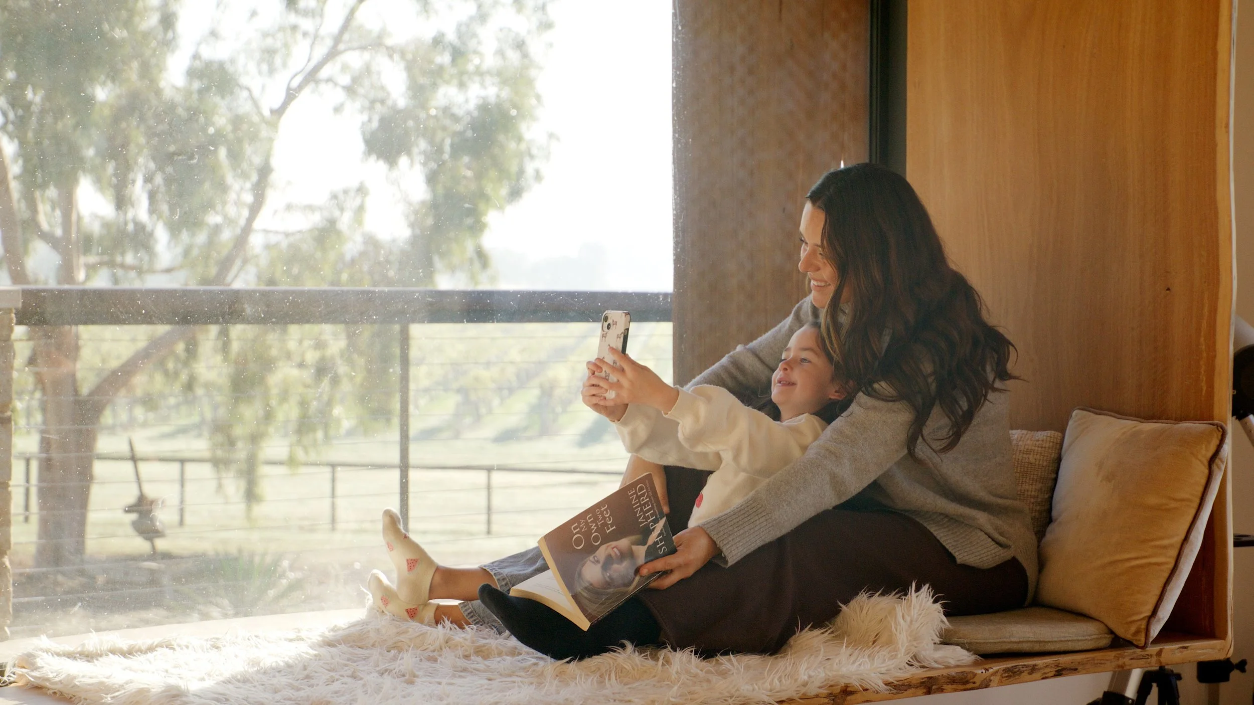 A woman and a young girl sitting together on a cozy bench by a large window, smiling and taking a selfie. The girl holds a magazine and is leaning on the woman, who is smiling. Natural light illuminates the scene, with a view of trees outside.