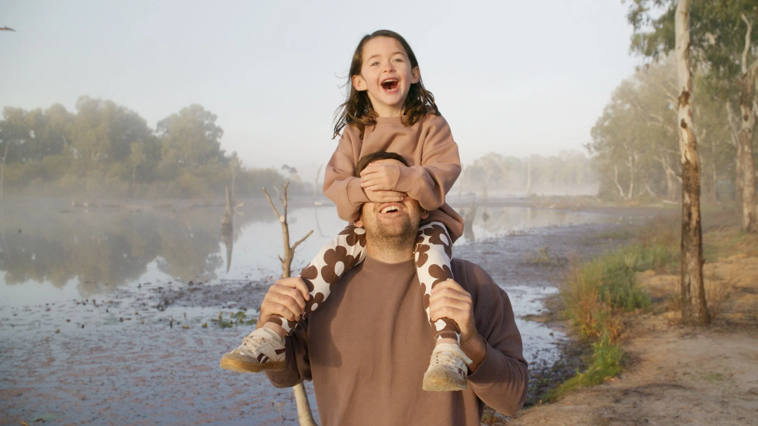 A man carrying a young girl on his shoulders outdoors near a river, with trees and misty weather in the background.