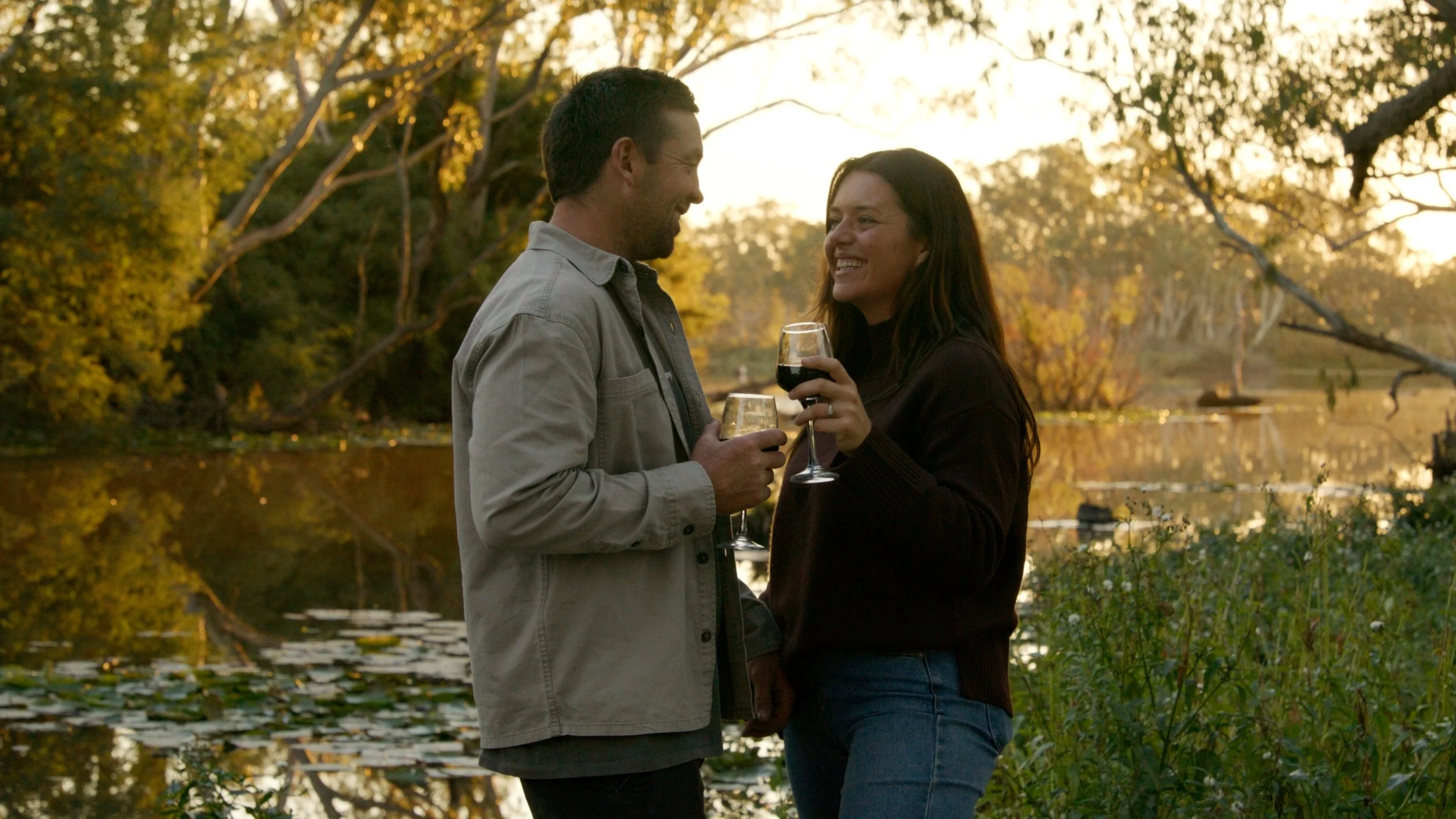 A man and woman enjoying a sunset by the water while holding glasses of red wine, smiling at each other.
