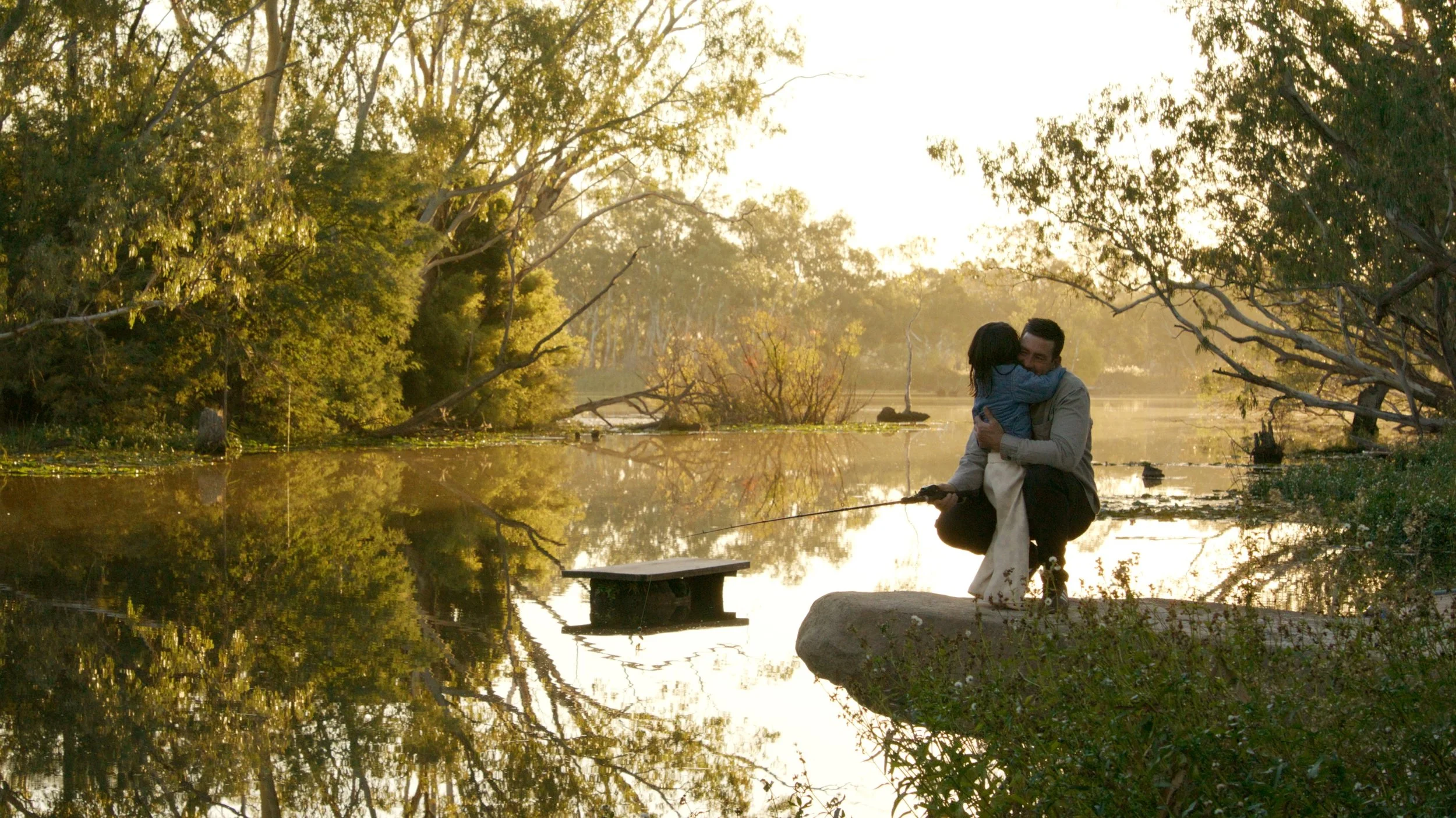 A man and a woman share an embrace on a dock beside a calm river surrounded by trees, with sunlight filtering through the branches.