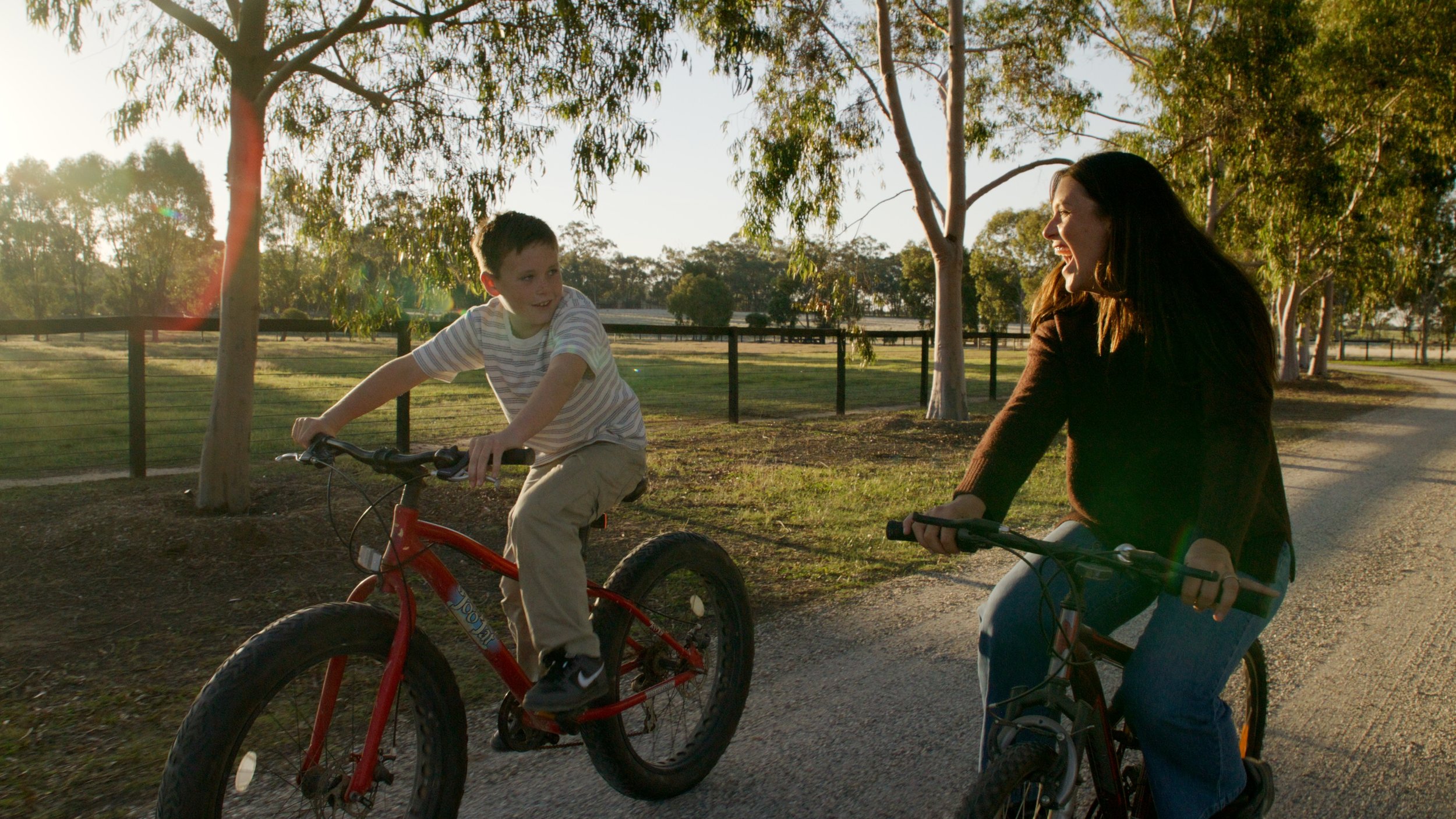 A woman and a boy riding bikes and smiling on a sunny day in a park with trees and a fence in the background.