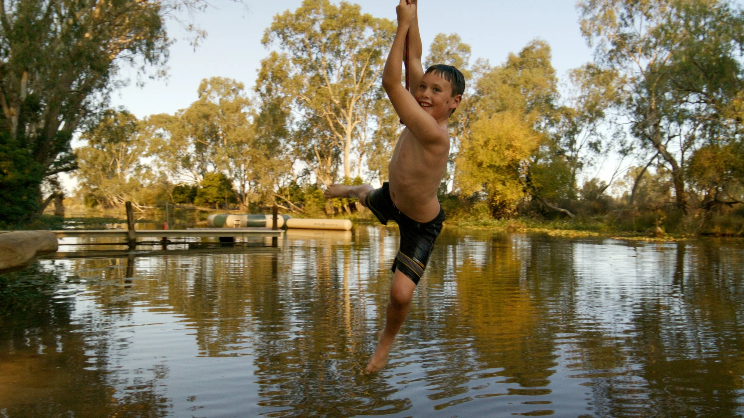 A young boy with dark hair smiling and hanging from a tree branch over a body of water, wearing dark swim trunks, with a background of trees and a dock.