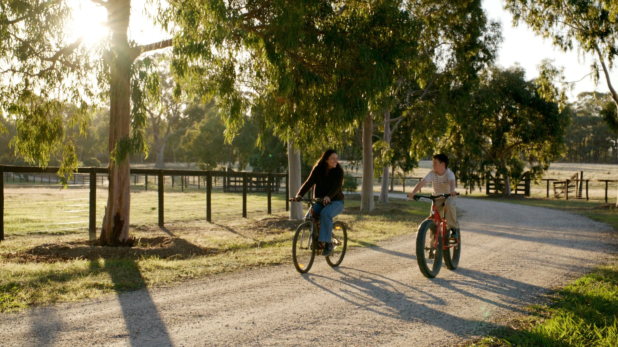 A woman and a boy are riding bicycles along a dirt path in a park during late afternoon or early evening, with sunlight filtering through trees, creating long shadows.