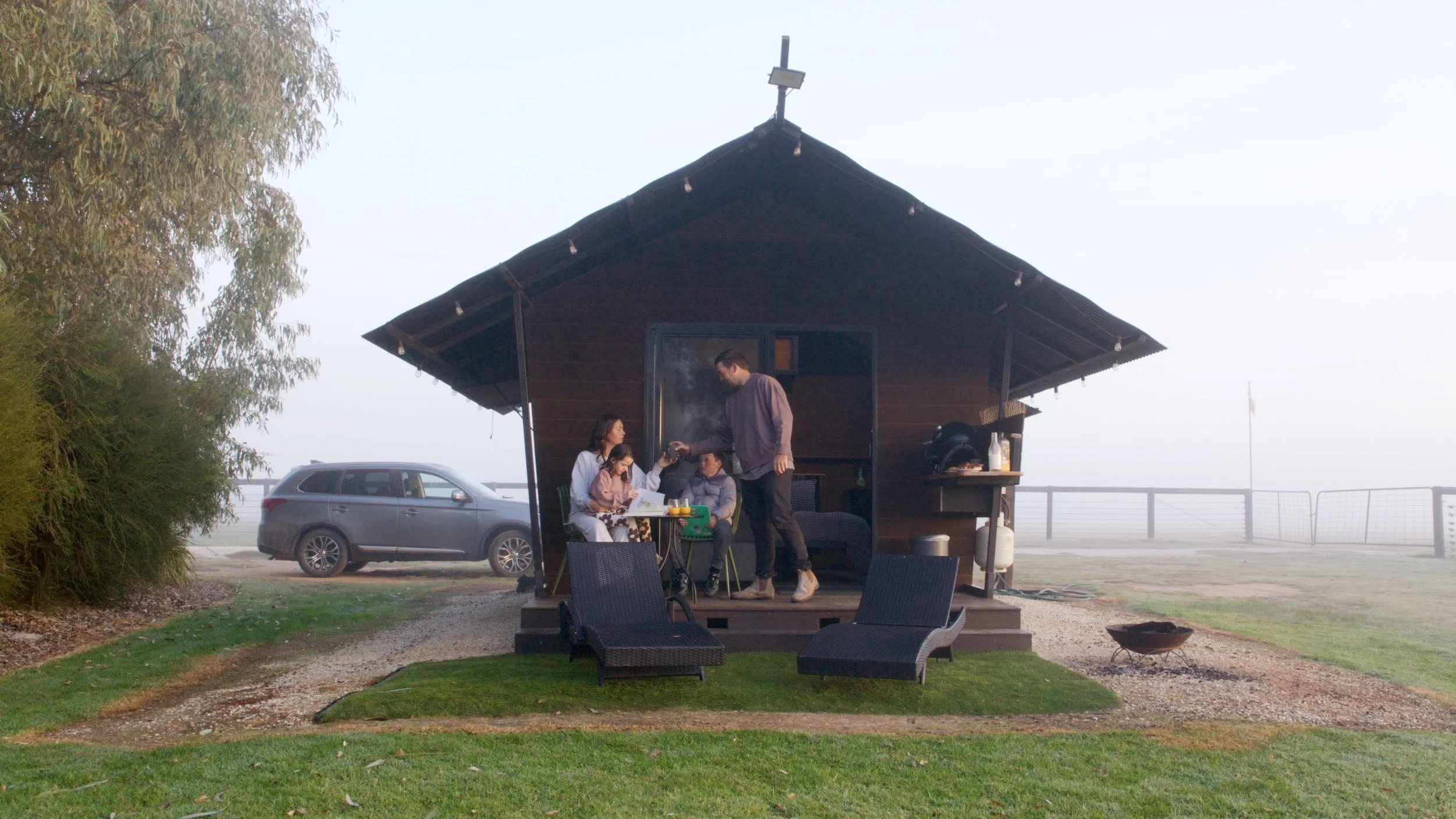 A family is gathered on the porch of a small wooden house with a car parked nearby. The scene appears foggy or misty, with trees and a fence in the background.
