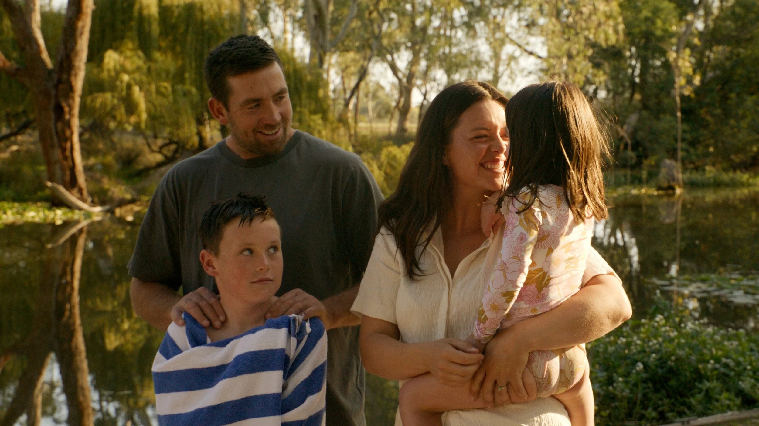 A family of four enjoying a moment outdoors near a pond with trees in the background during sunset, with the mother holding a young girl and the father and son standing beside them.