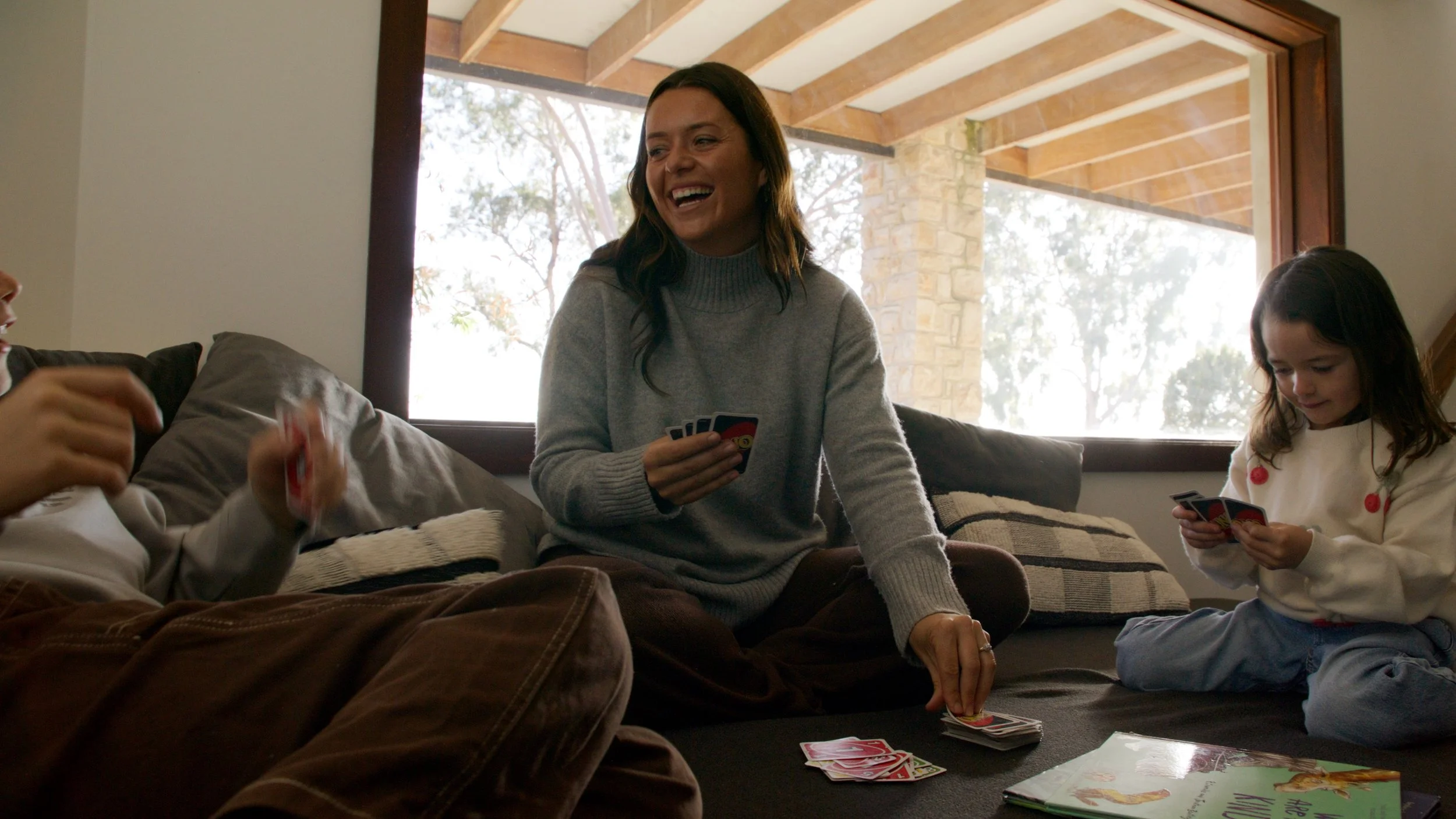 A woman and two children playing card games on a couch in a sunlit living room.