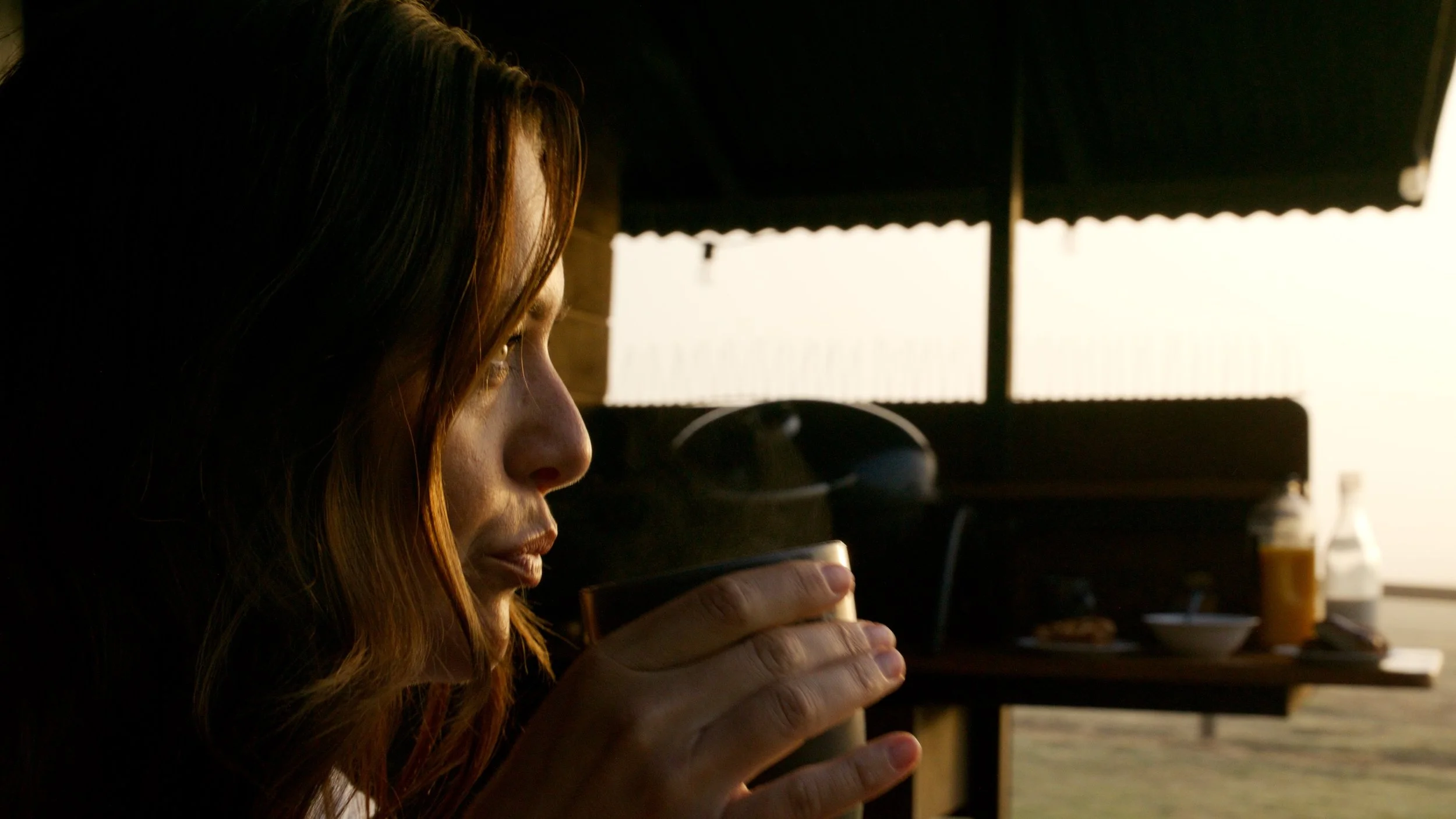 A woman with reddish-brown hair sipping from a glass in a kitchen with a dark window shade and kitchen items in the background.