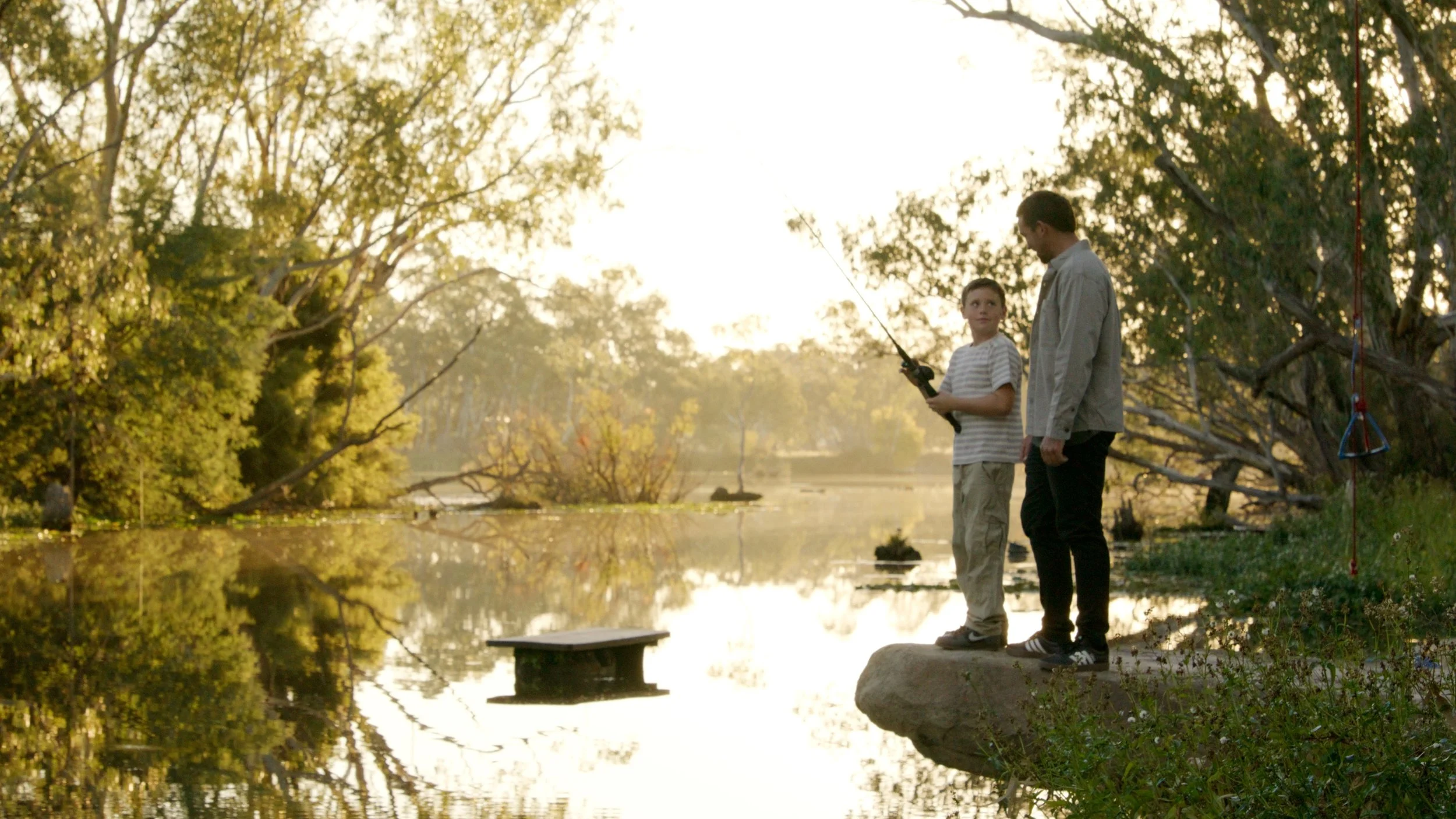 A man and a boy fishing by a river at sunset, standing on a large rock surrounded by trees.