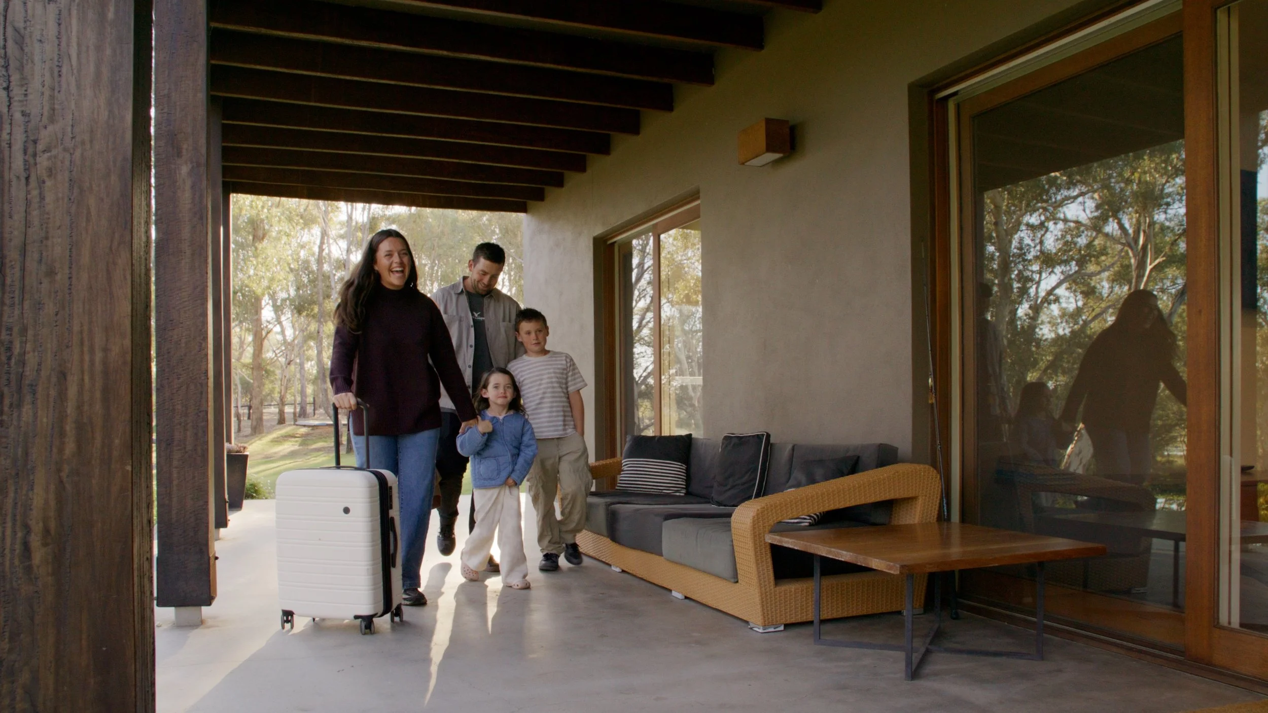 Family arriving at a home, carrying luggage, with sunlight and trees outside.