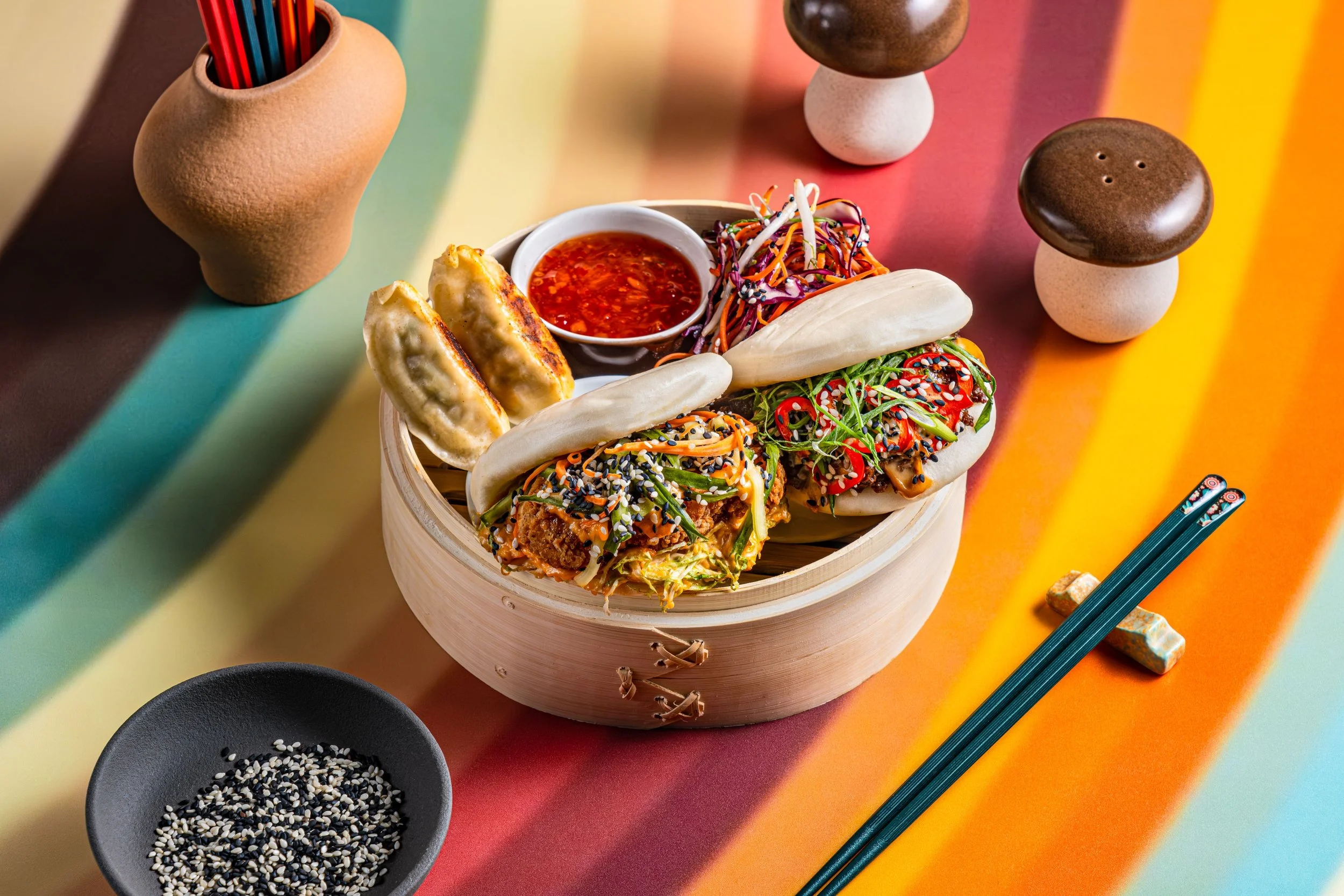 Assorted bao buns and dumplings with dipping sauce on colorful background, sesame seeds in a small dish, chopsticks, and mushroom-shaped salt and pepper shakers.