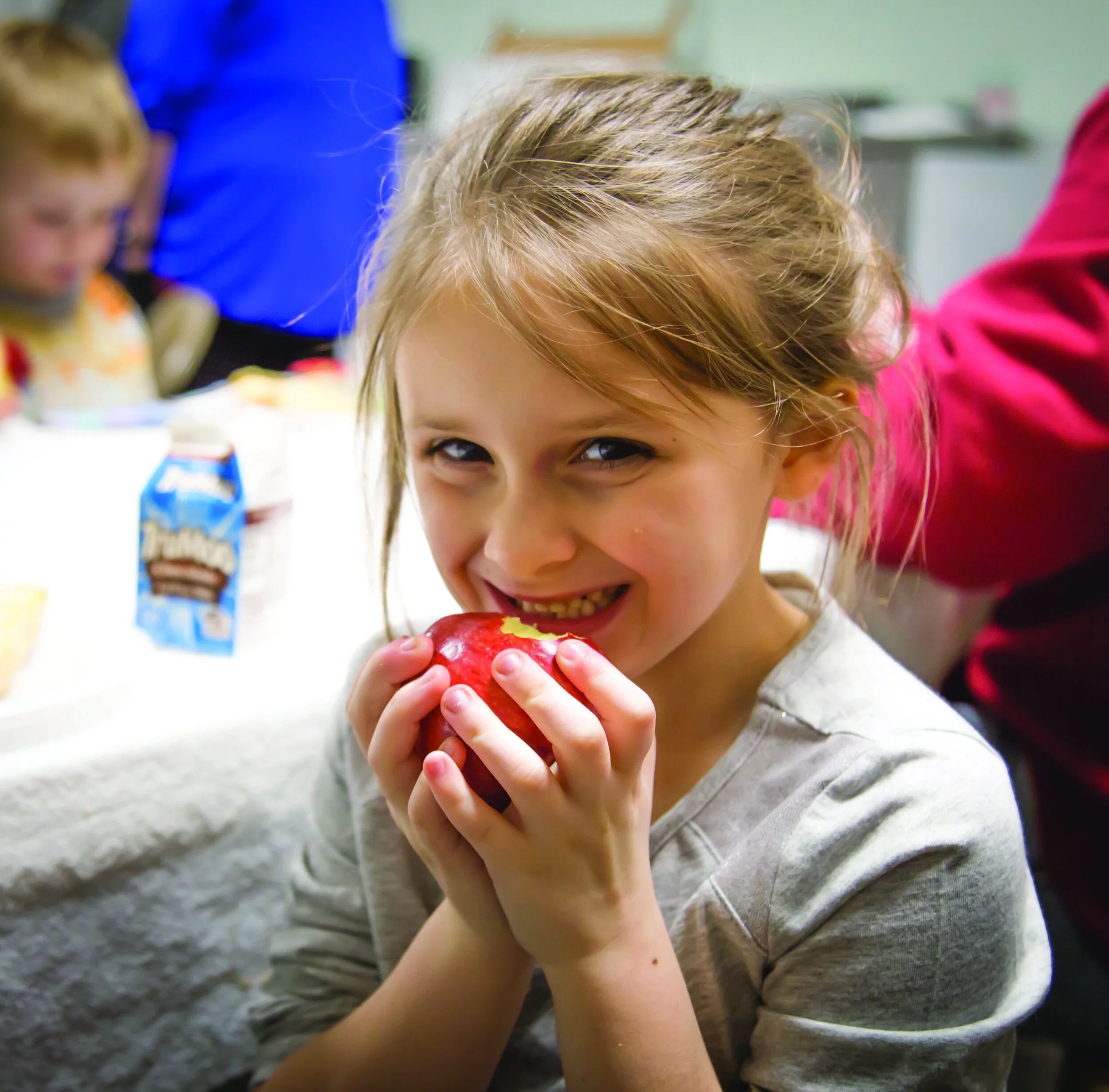 A young girl with light brown hair smiling and holding a red apple near her mouth at a gathering, with other children and food in the background.