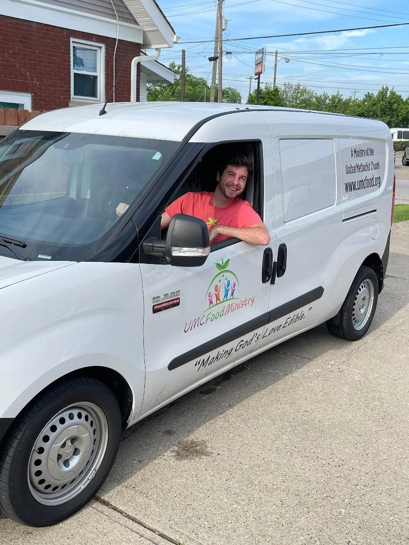 A smiling man sitting in the driver's seat of a white delivery van parked on a street. The van has logos and text for UMC Food Ministry, including a colorful logo with three figures and a green leaf, and phrases like 'Making God's Love Edible' and a website link.