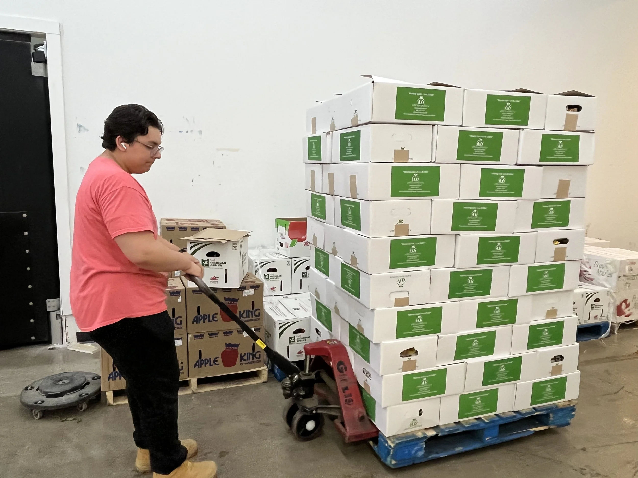 Person in a pink shirt and black pants handling a box on a pallet jack near large stacks of produce boxes in a warehouse.