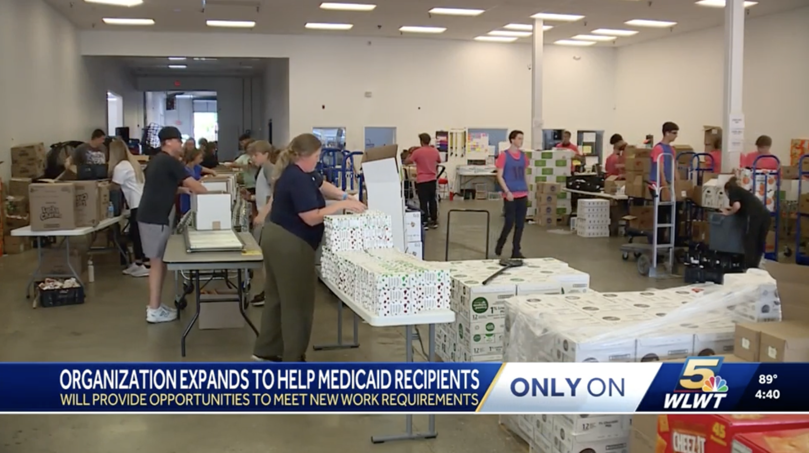 People working in a large room with tables and boxes, packing supplies, likely involved in assembling or distributing packages, with a news ticker at the bottom about an organization helping Medicaid recipients.