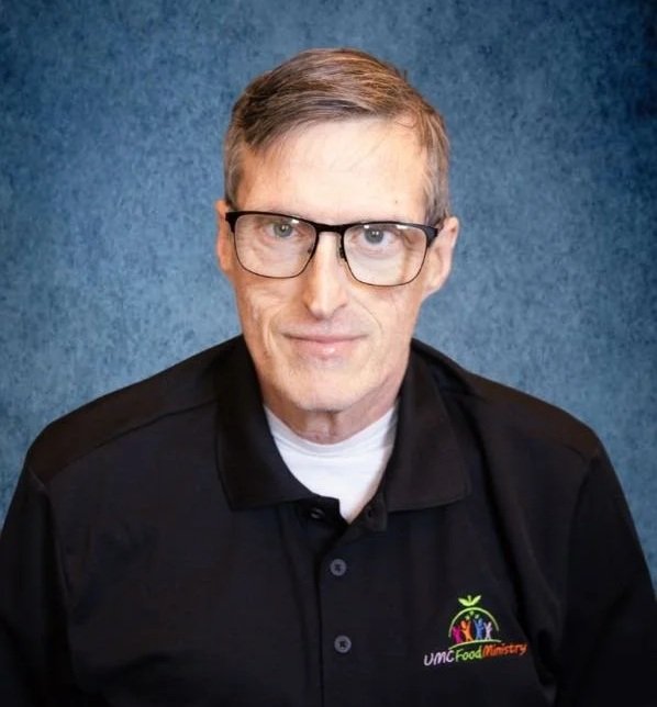 A man with short, light brown hair and glasses, wearing a black polo shirt with the logo 'UMC Food Ministry' on it, standing in front of a blue textured background.