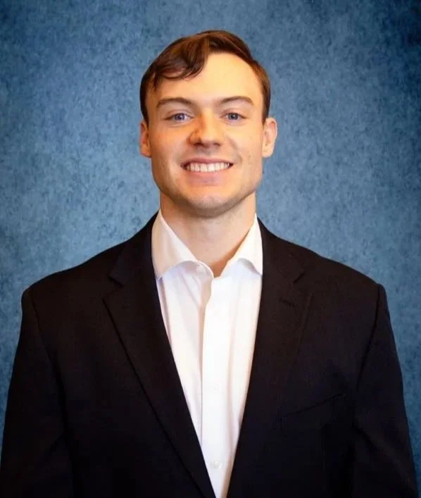 Young man with short brown hair smiling, dressed in a black suit and white shirt, standing against a blue textured background.