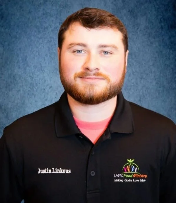 A young man with light skin, short brown hair, and a beard, wearing a black polo shirt with a logo and text on it, standing against a textured blue background.