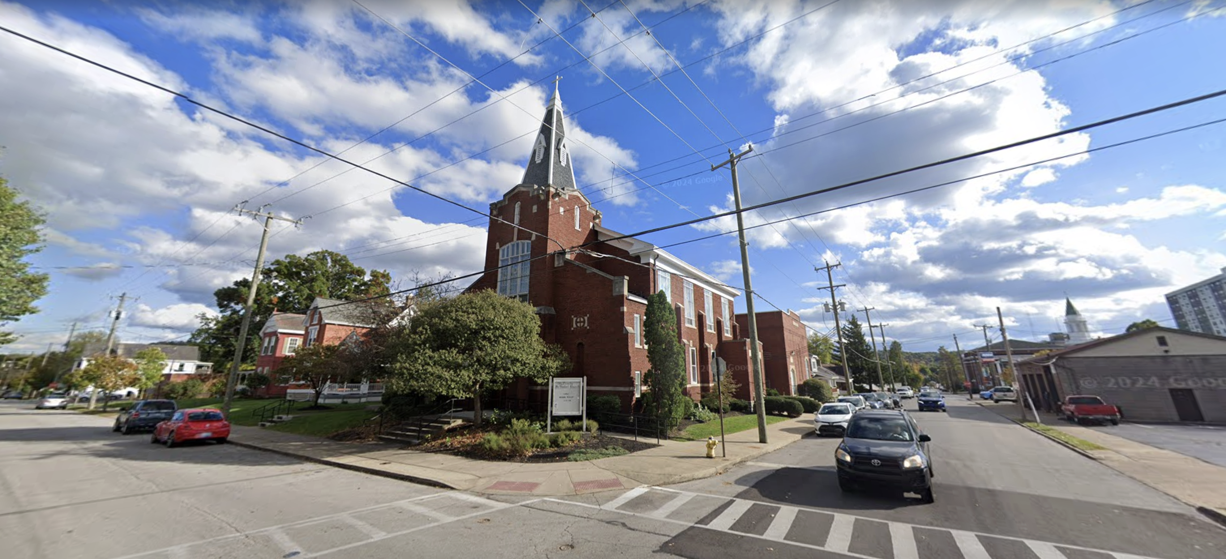 A red brick church with a tall steeple under a partly cloudy blue sky, surrounded by trees, parked cars, and a street in a small town.