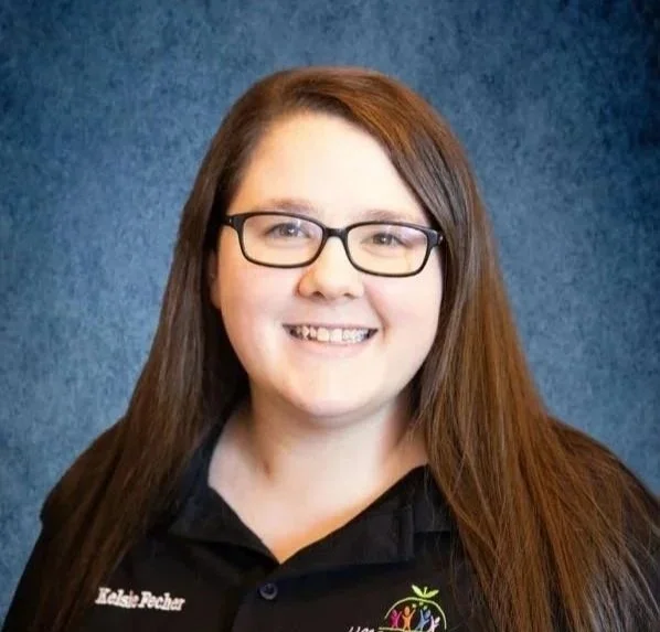 A woman with long brown hair and glasses smiling, wearing a black polo shirt with logos and text, standing against a blue textured background.