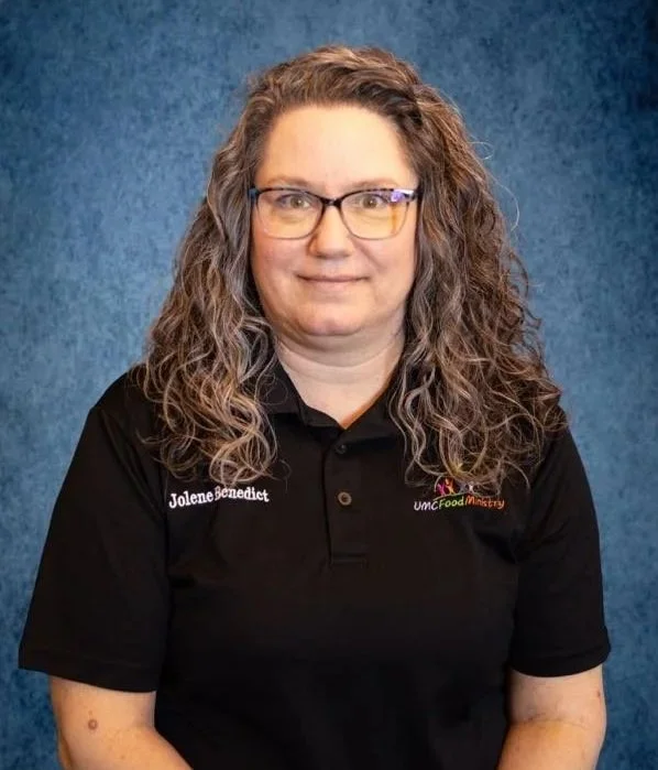 A woman with curly hair, glasses, wearing a black polo shirt with an embroidered name tag that reads 'Jolene Benedict' and a logo for 'UMC Food Ministry.' She is standing against a blue textured background.