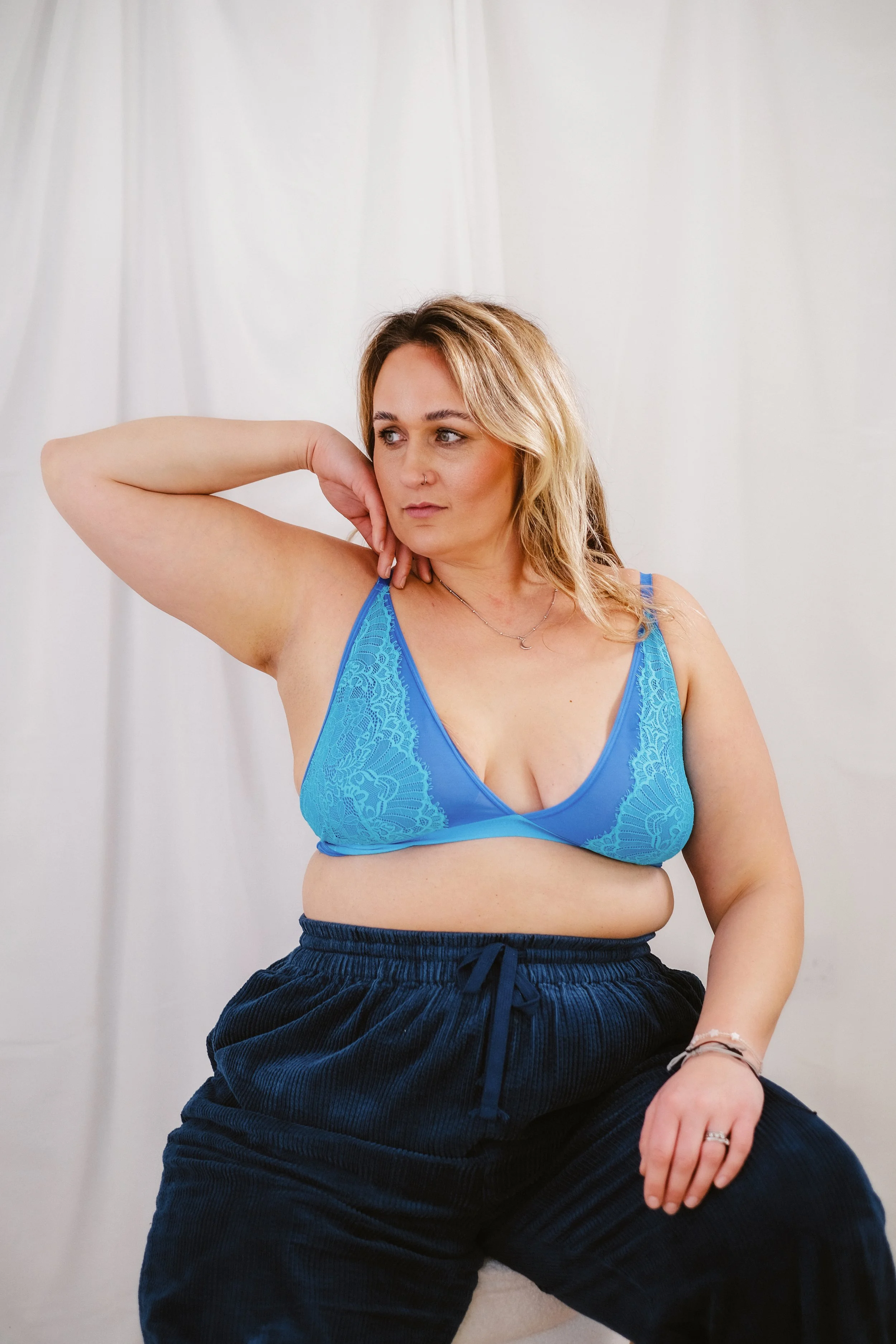 Black and white photo of a woman in a black bra and lace underwear, with one arm raised above her head, posed against a plain wall. Perfect photoshoot. Body Positive