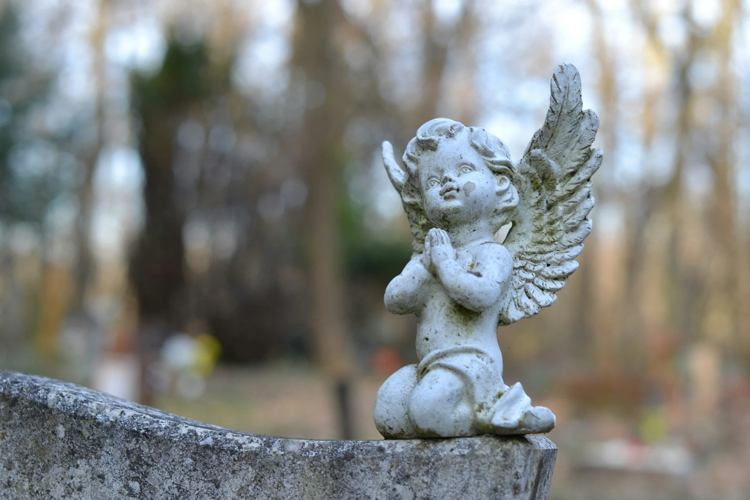 A small angel statue with wings, kneeling with hands pressed together in prayer, on a stone surface in a blurred outdoor background.
