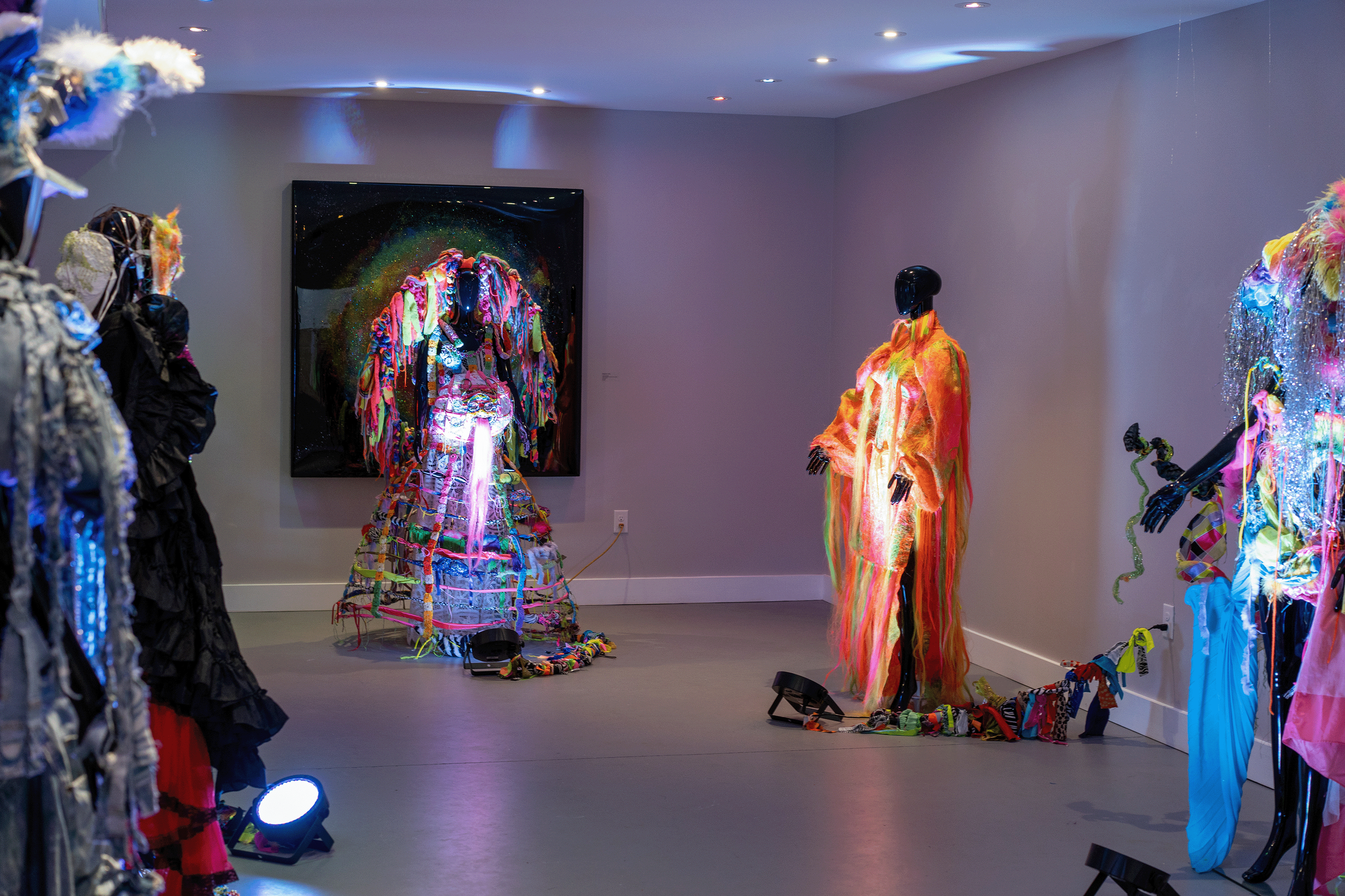 Art exhibition featuring mannequins dressed in vibrant, colorful garments with various fabrics and textures, displayed in a gallery setting with spotlights.