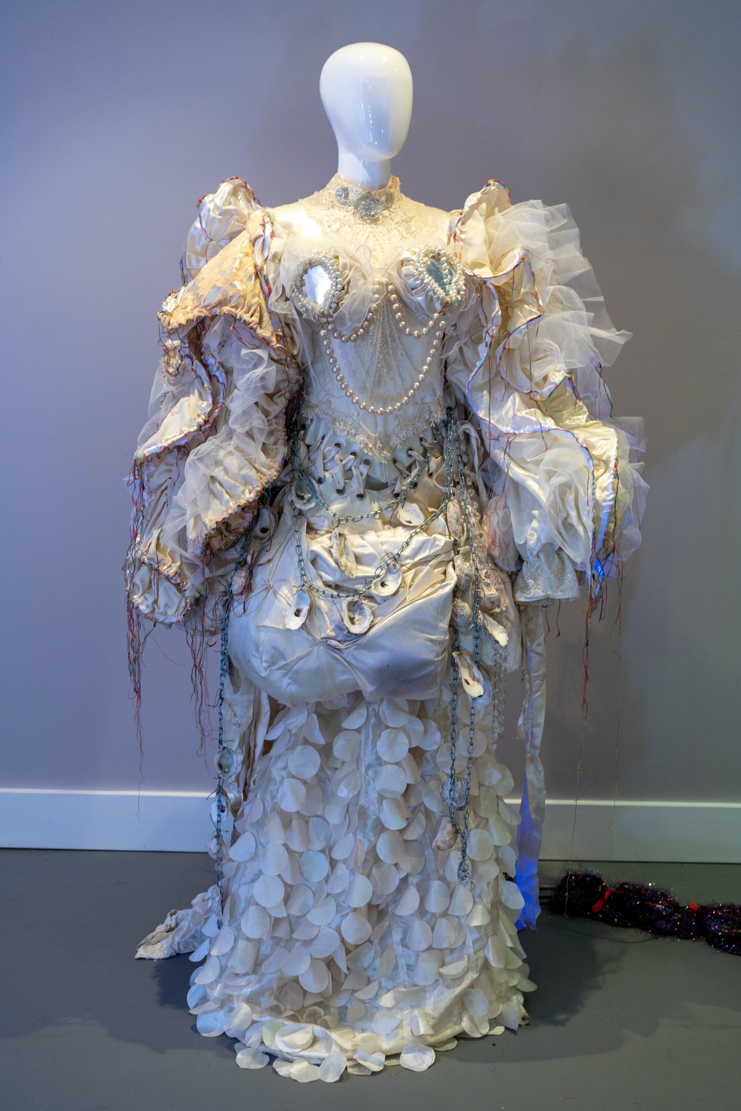 Mannequin displaying an elaborate, decorative costume with ruffled sleeves, pearl embellishments, and textured fabric, set against a plain background.