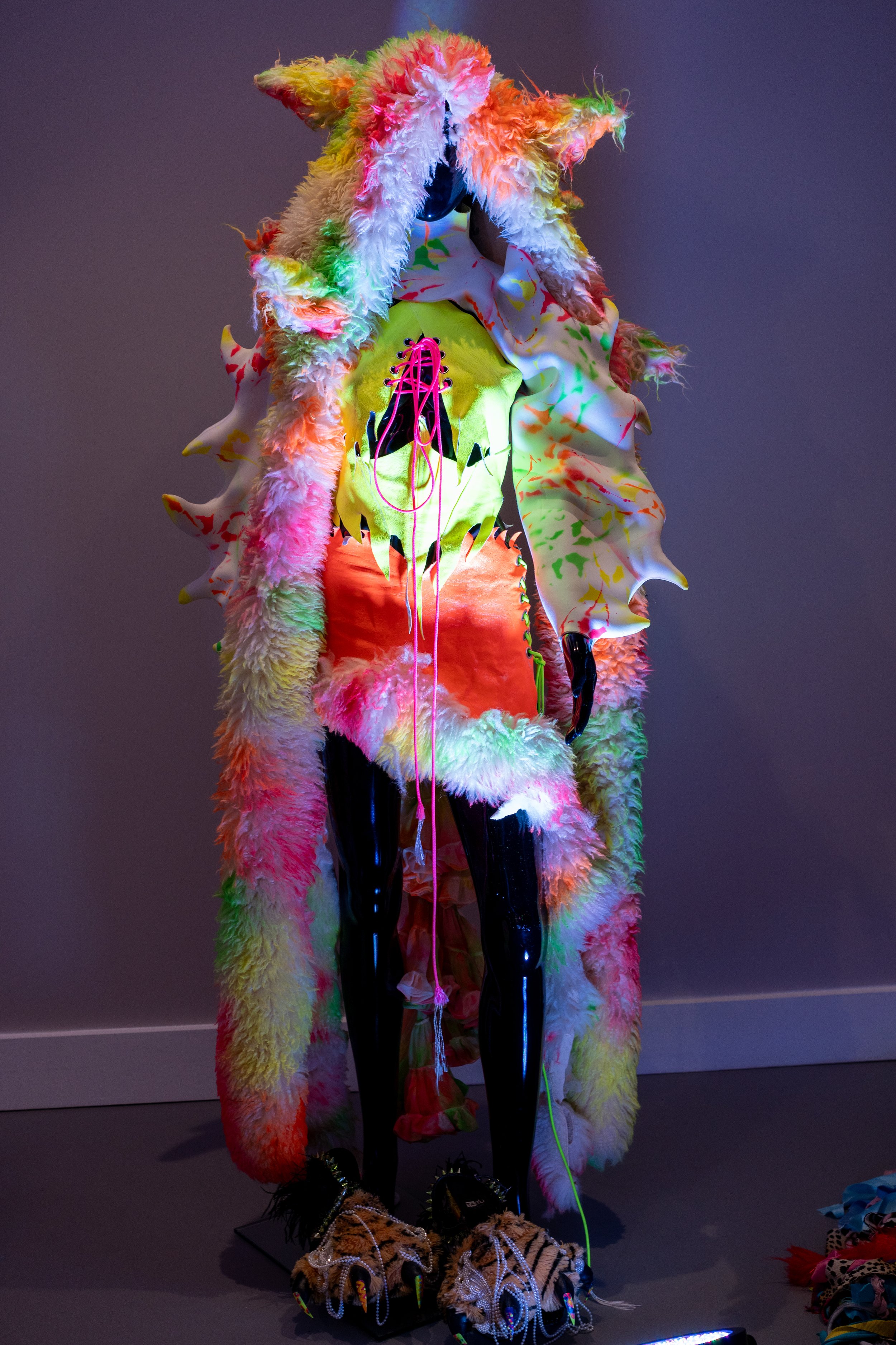 Mannequin wearing a colorful, furry costume with neon accents, resembling a fantasy creature. The outfit includes a shaggy, multicolored headdress with spiky protrusions, a neon yellow and orange torso, and unique oversized shoes.