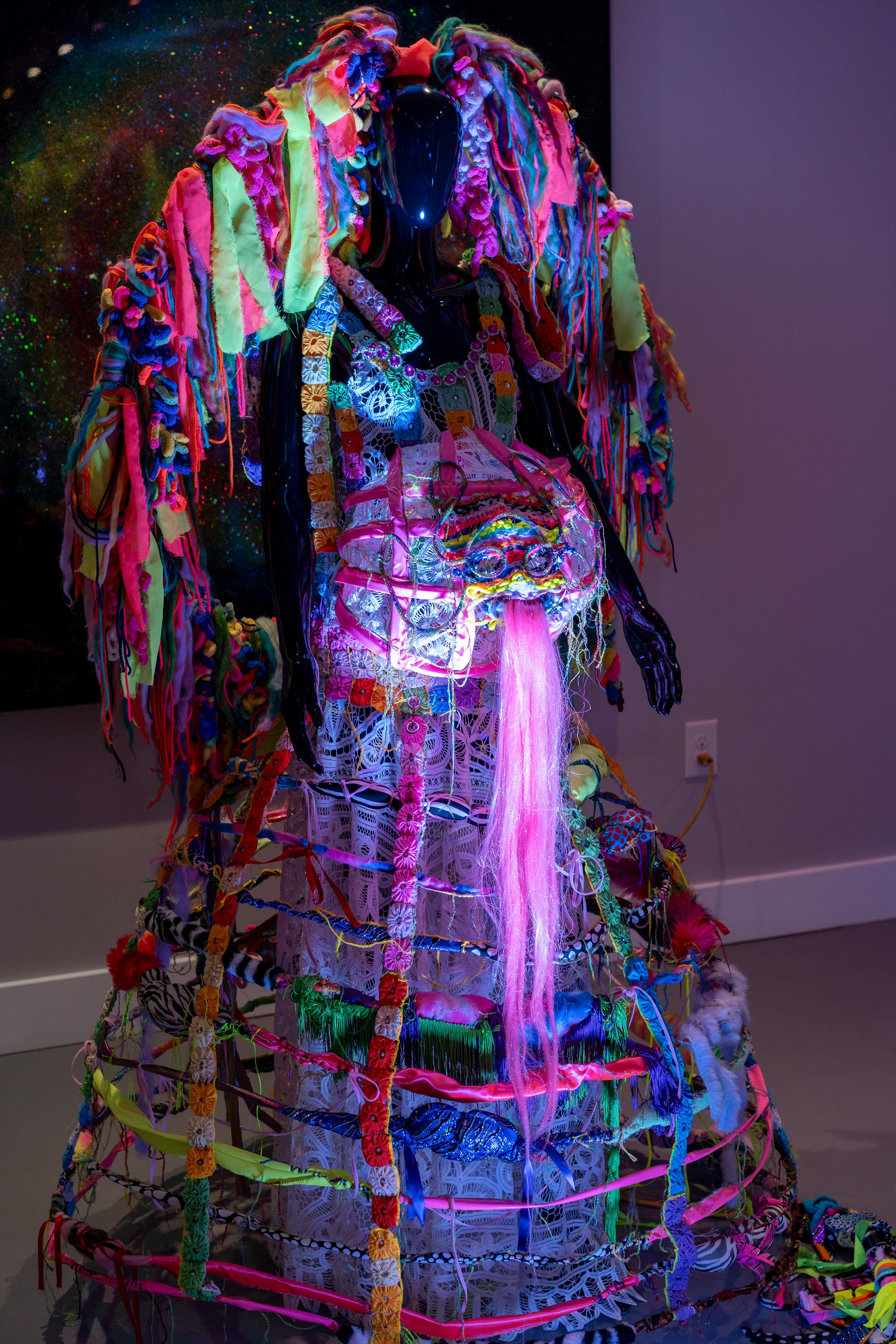 Colorful mannequin art piece adorned with vibrant fabric strips and yarn, featuring a black mannequin form, elaborate textile design, and intricate details against a dark background. Neon and pastel hues dominate the artwork.