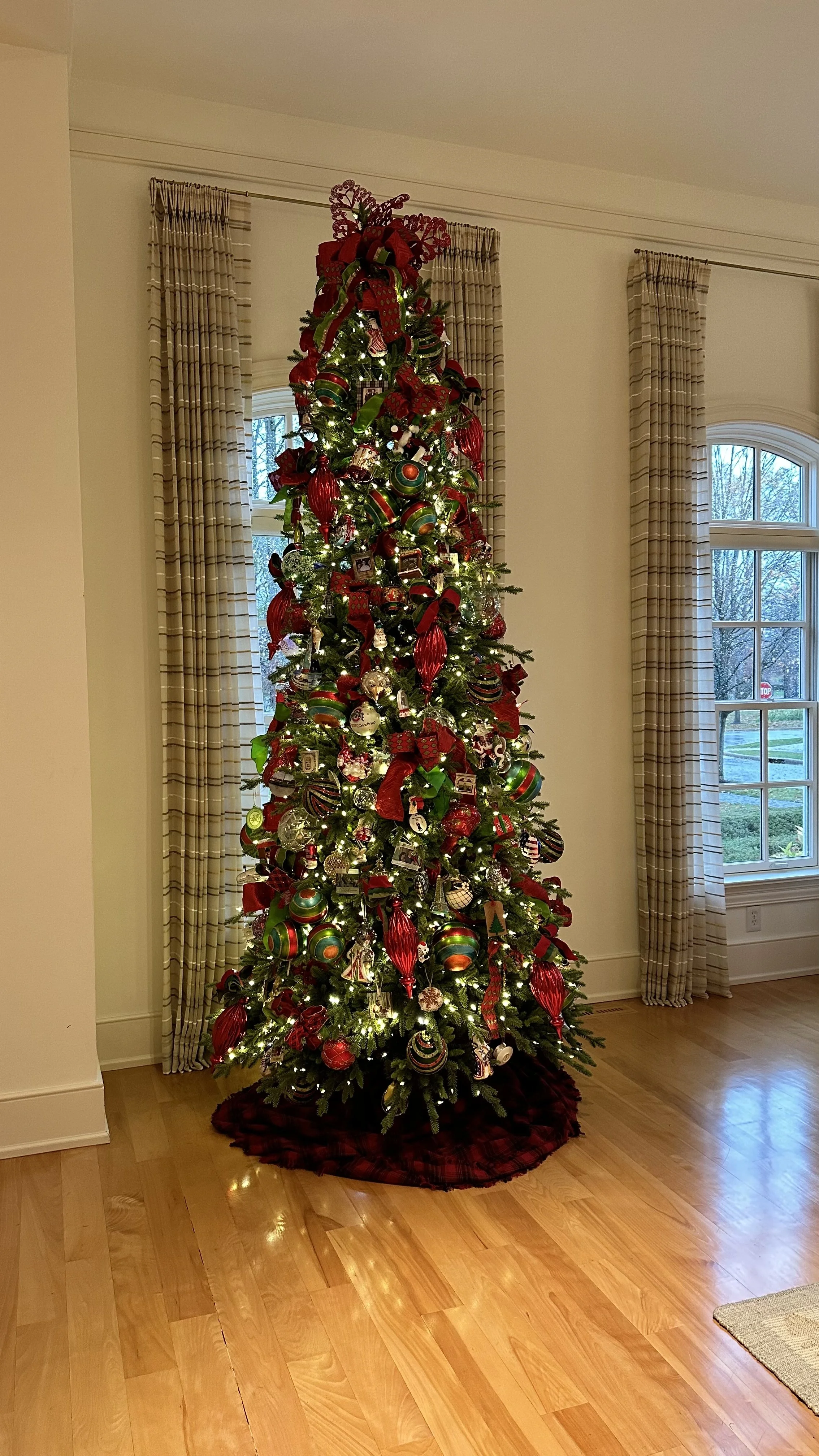 Decorated Christmas tree with red, green, and gold ornaments and ribbons in a living room with hardwood floors and large windows.