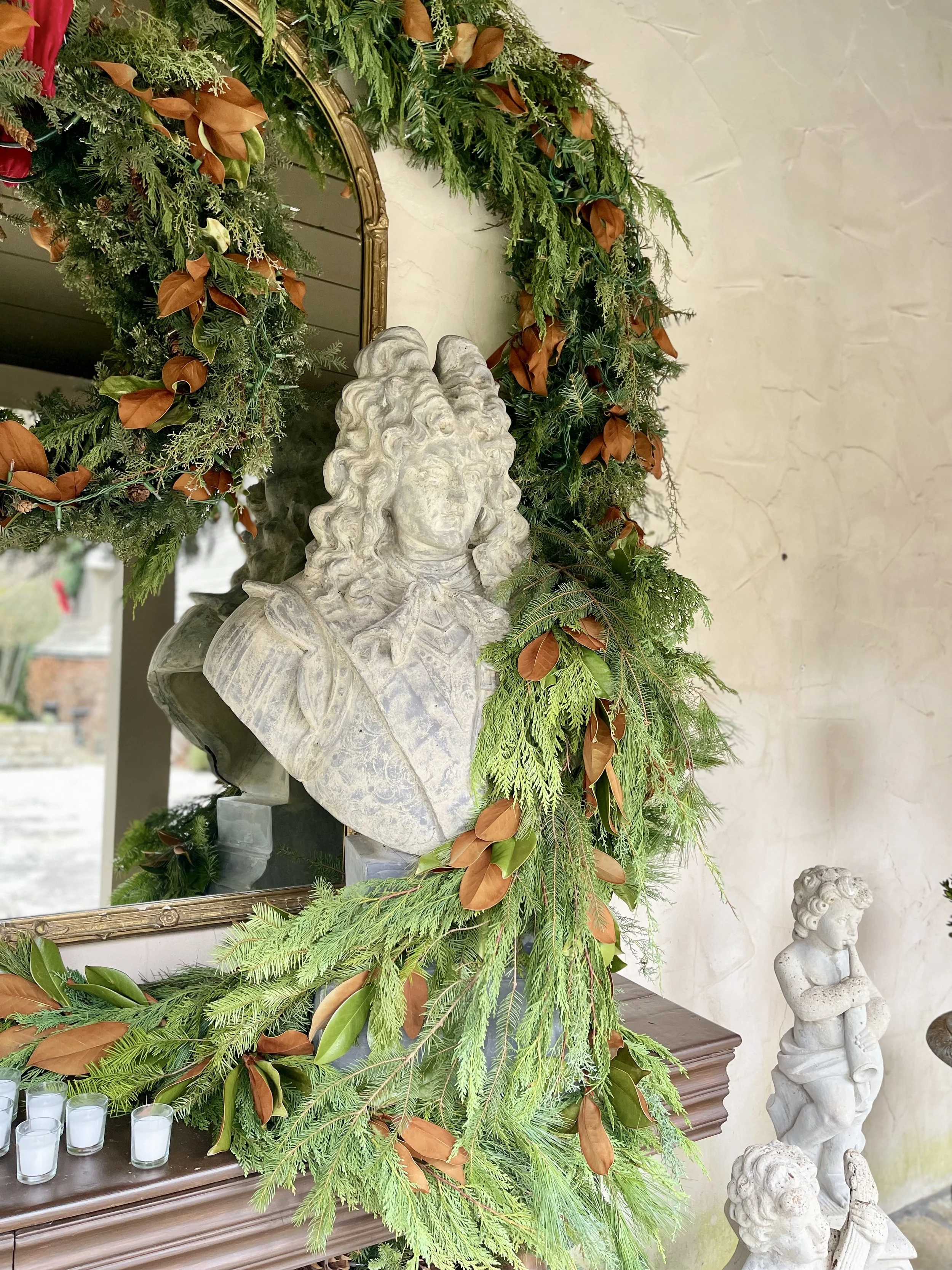 Decorative display featuring classical stone busts, surrounded by a lush green and brown garland with a mirror reflecting part of the arrangement and a beige textured wall in the background.