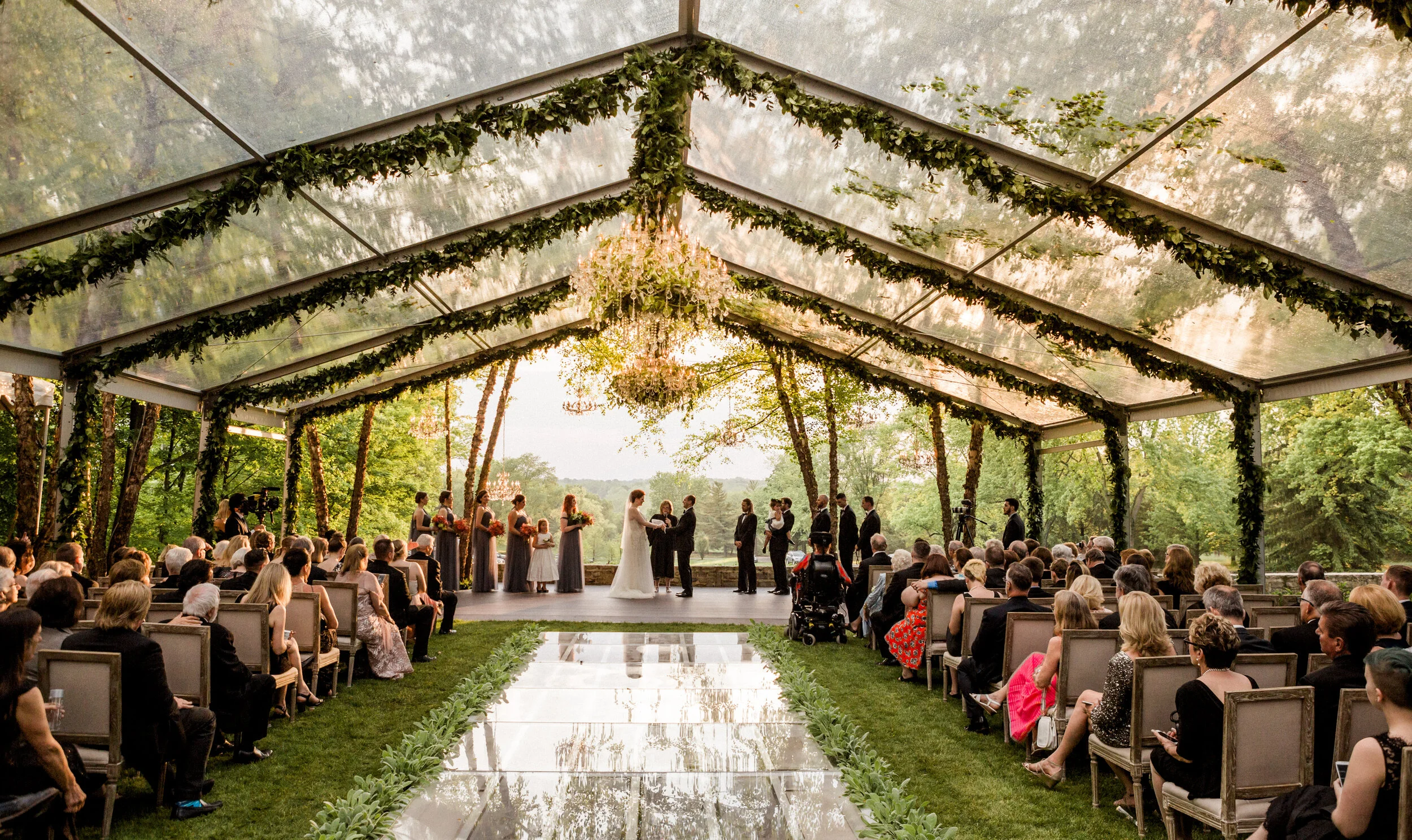 Outdoor wedding ceremony under a clear tent with floral decorations, featuring guests seated on both sides of a reflective aisle.
