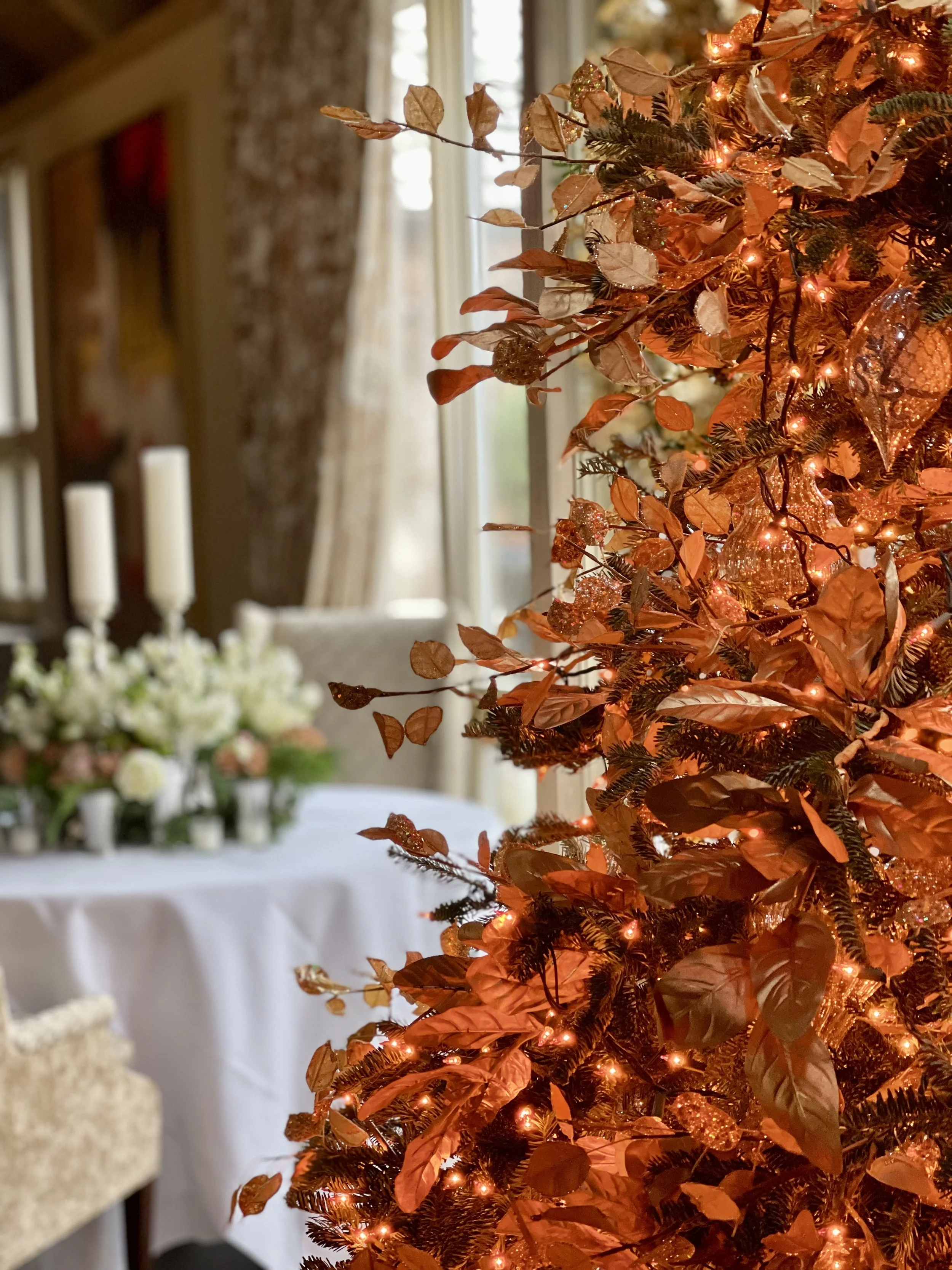 Close-up of a Christmas tree decorated with orange and gold leaves, ornaments, and lights, with a dining room background featuring a table with candles and floral arrangement.