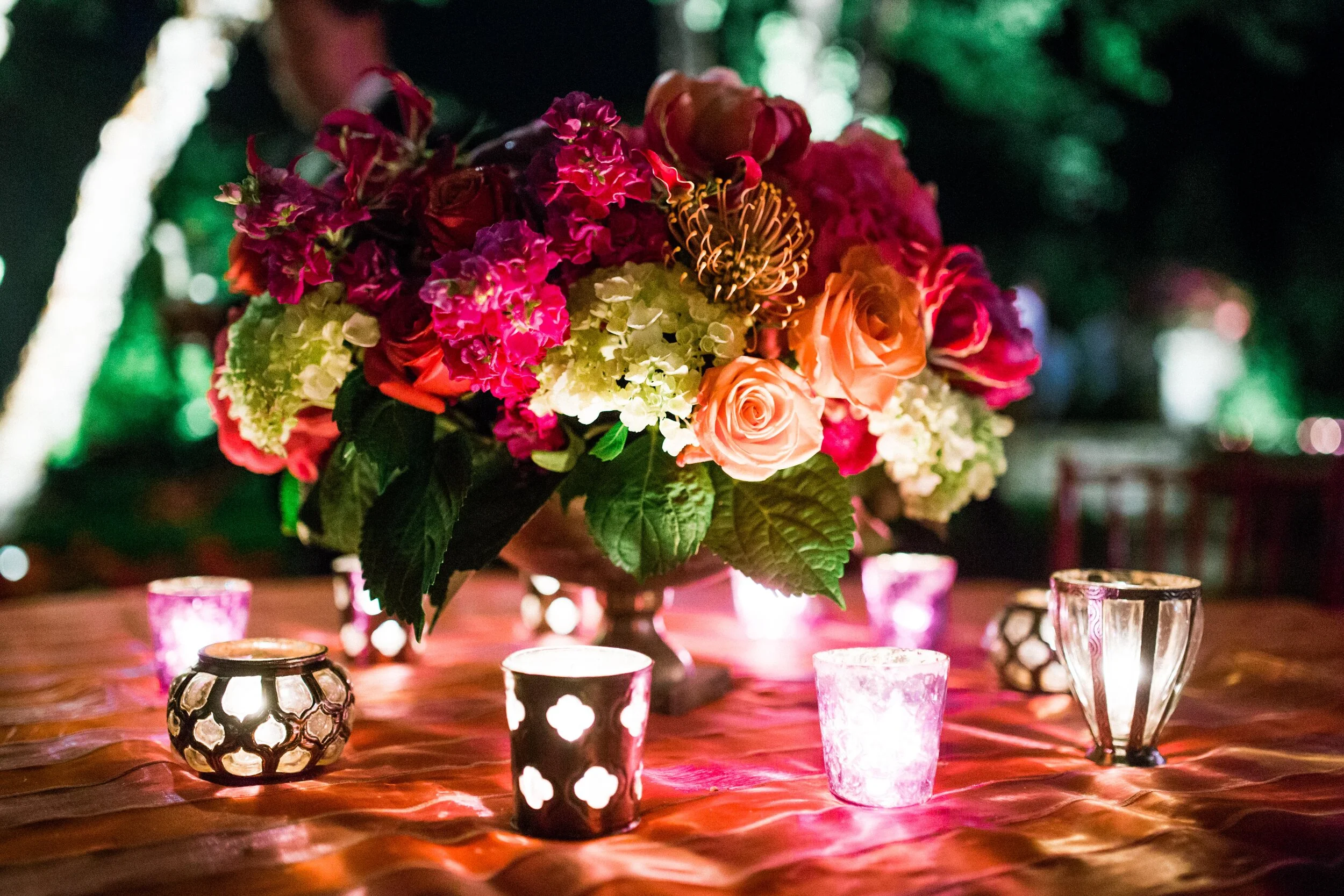 Colorful flower arrangement with roses, carnations, and leaves on table with glowing tealight candles in ornate holders.