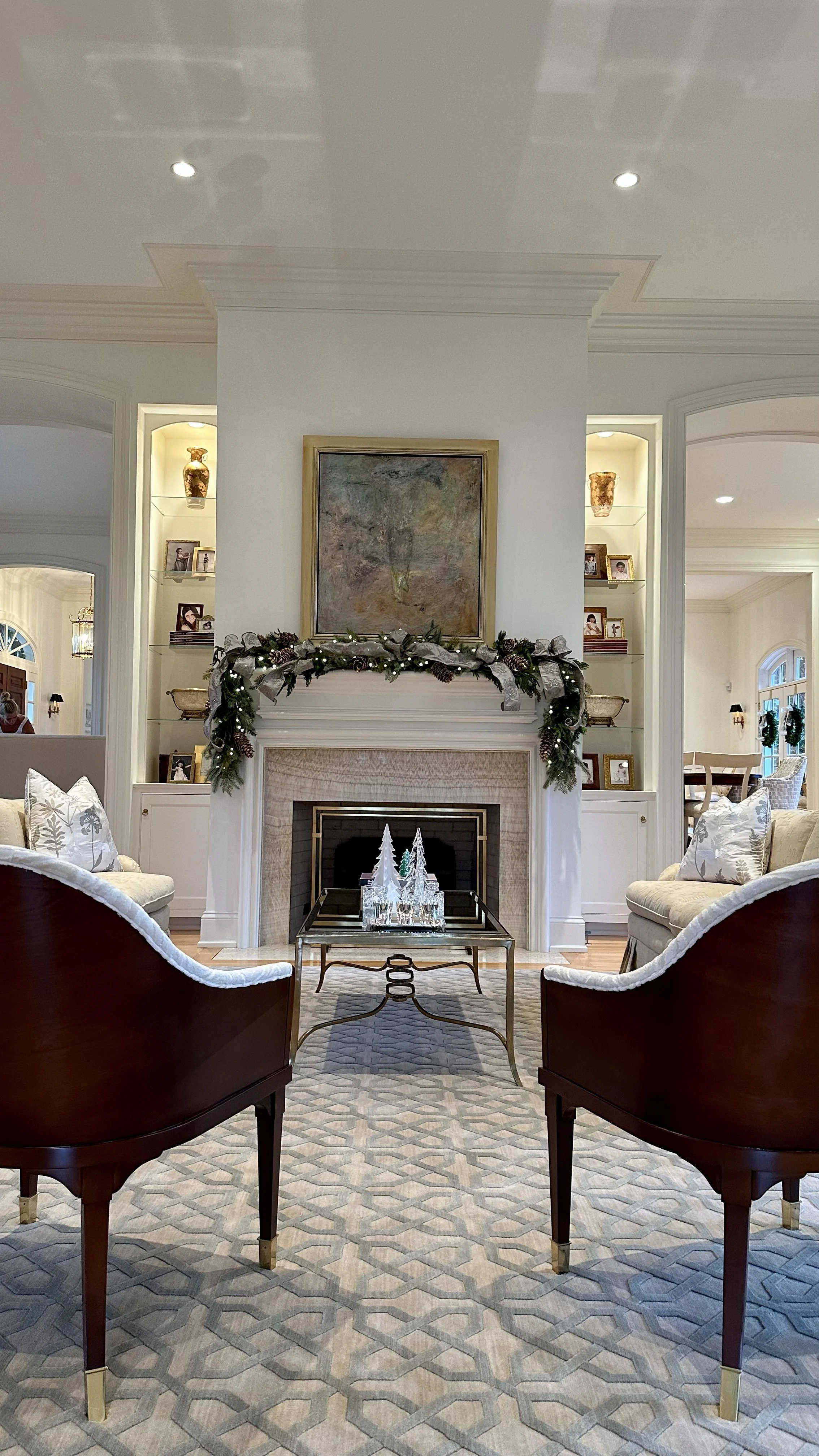 Living room with a fireplace decorated with a Christmas garland, framed artwork above, and glass Christmas trees on a coffee table, flanked by two cream-colored sofas and two dark wood armchairs.