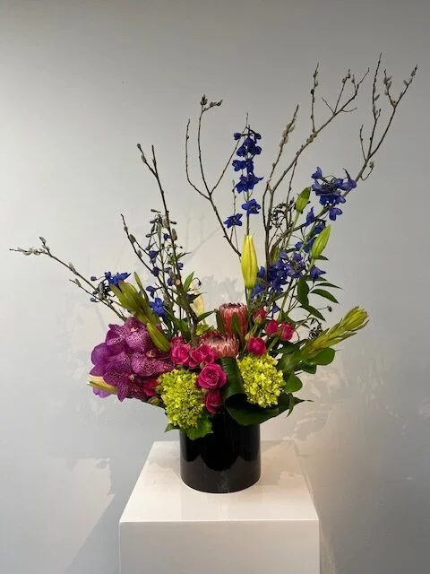 A mixed floral arrangement in a black vase featuring purple, pink, yellow, and blue flowers with green leaves and branches.