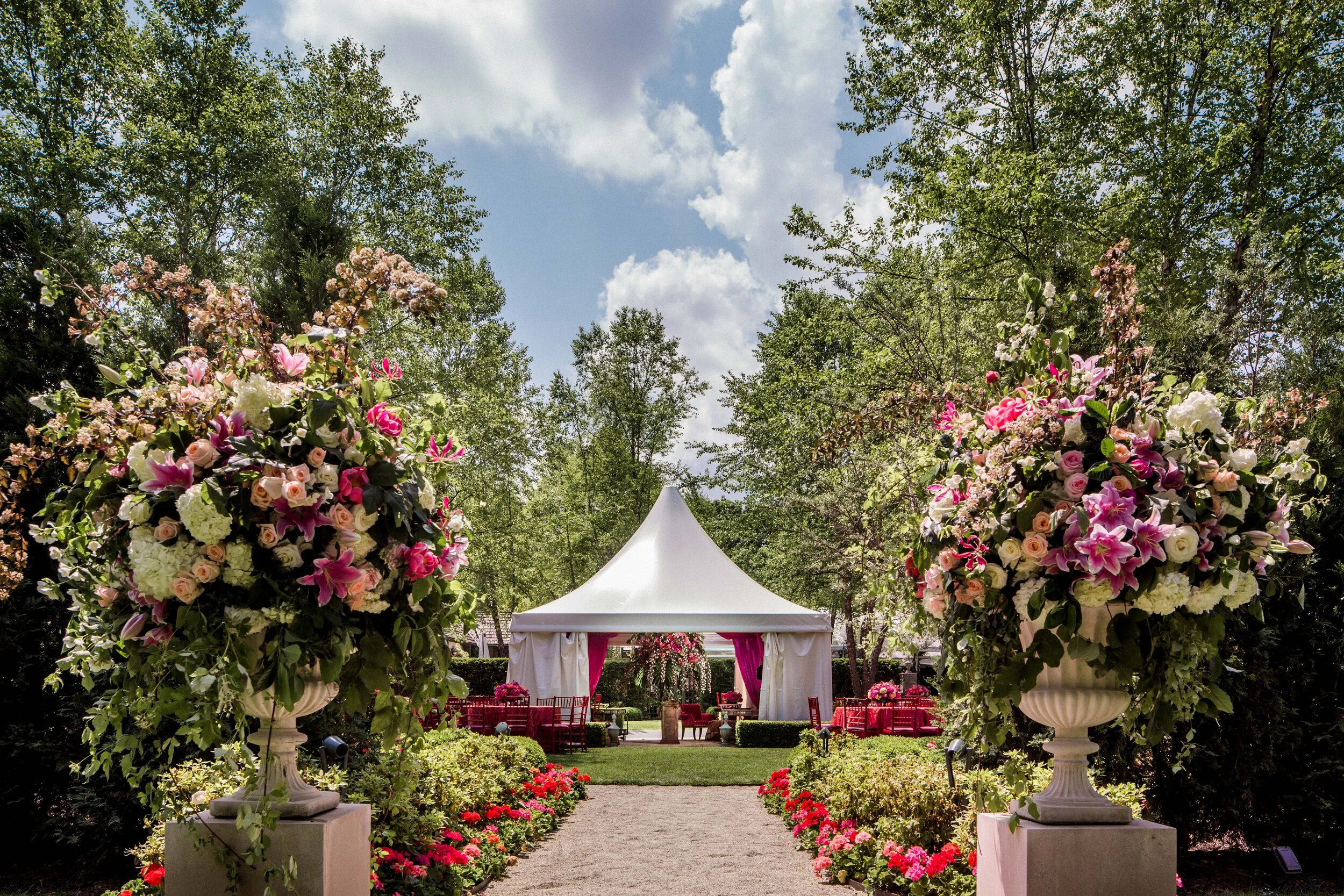 Outdoor garden wedding setup with a white tent, floral arrangements, red and pink roses, and green foliage along the aisle.