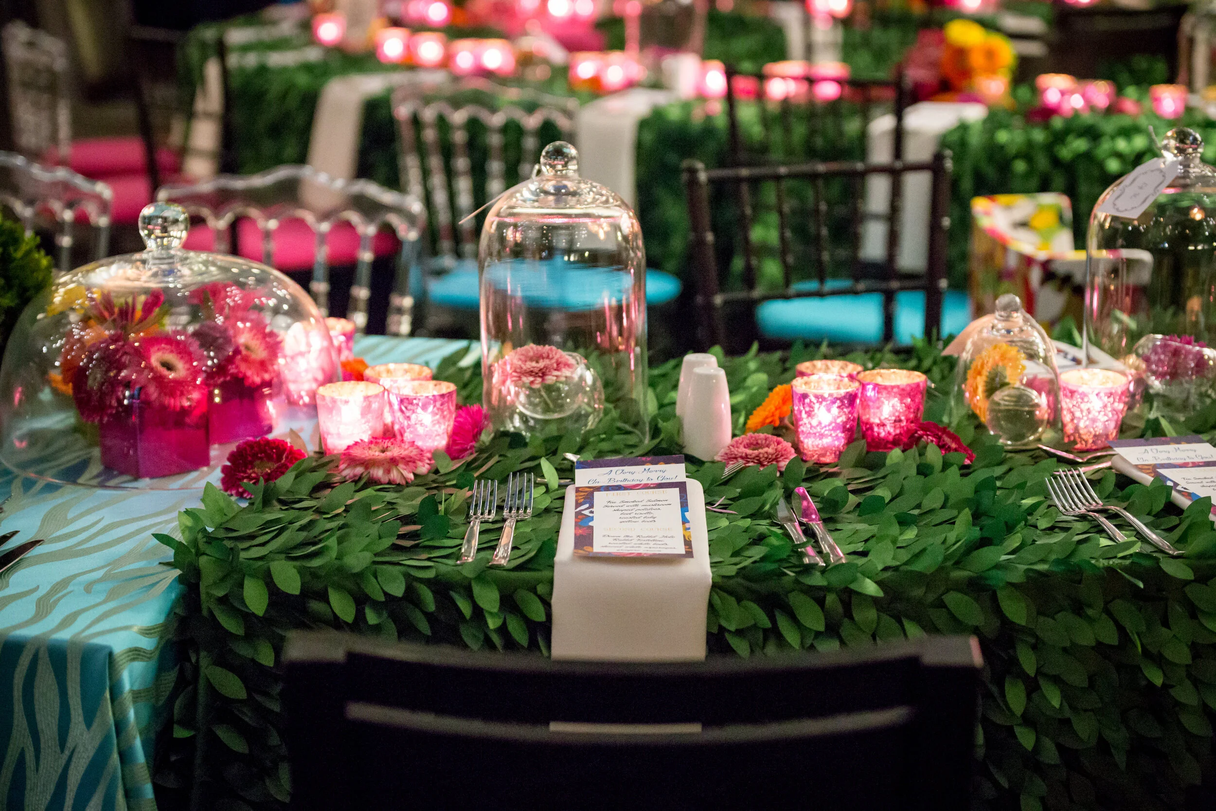 Decorated table setting with greenery tablecloth, pink floral arrangements under glass cloches, pink lit candles, and place settings with menus and cutlery.