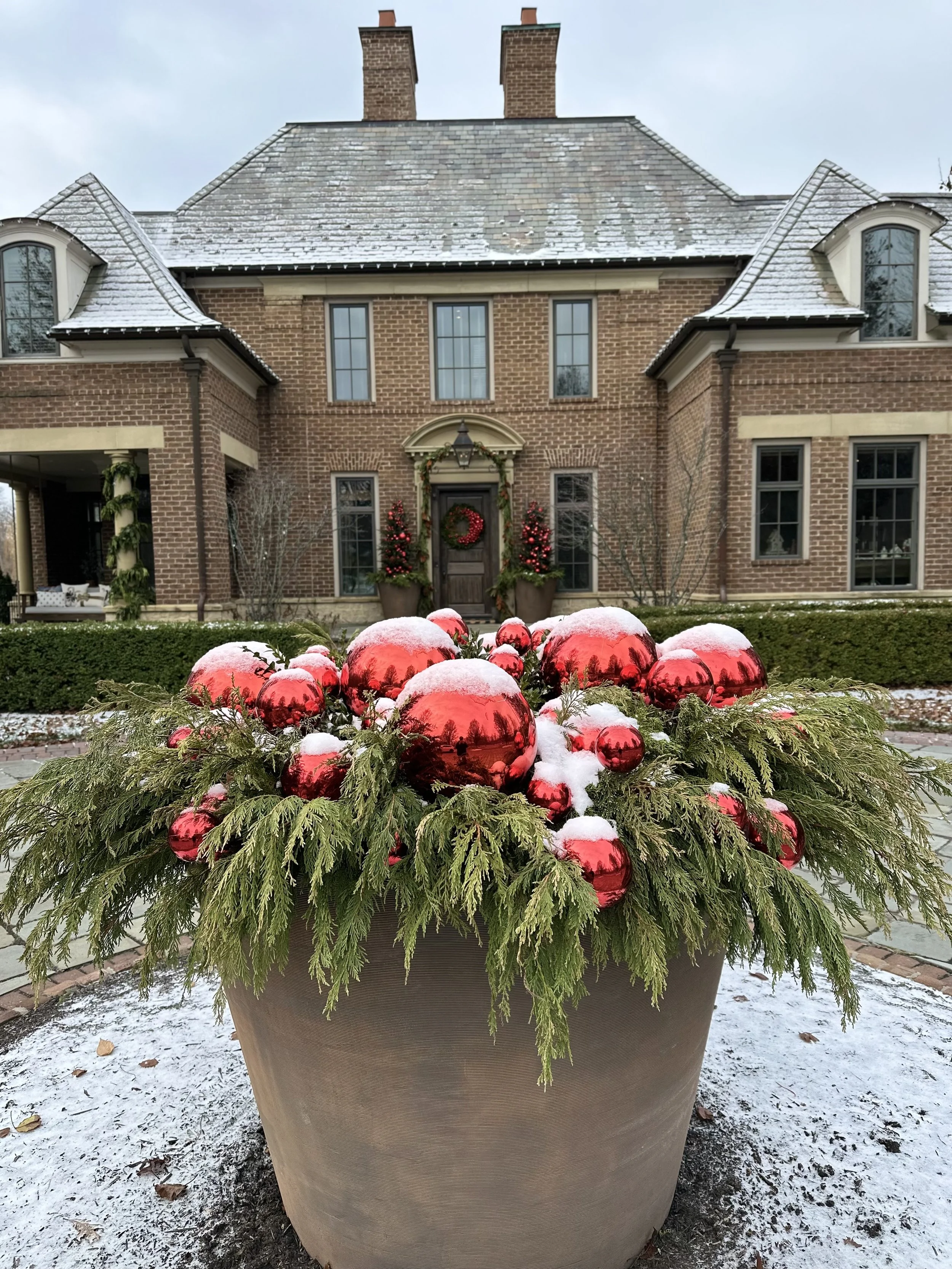 A large potted evergreen decoration with red Christmas ornaments and snow, in front of a brick house with holiday decorations on the door, during winter.
