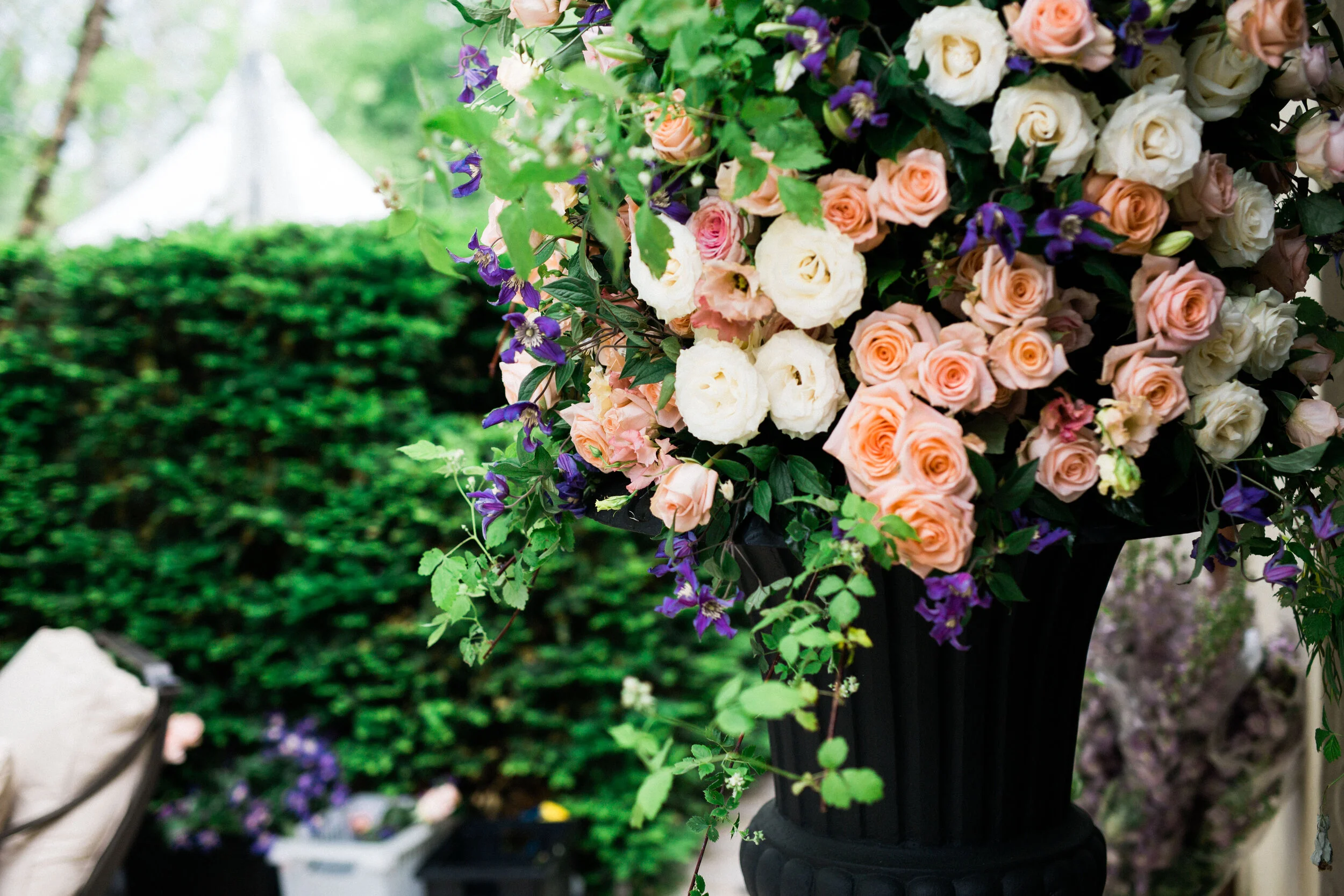 A large floral arrangement featuring peach and white roses with purple flowers and green foliage in a black vase, set against a garden background.