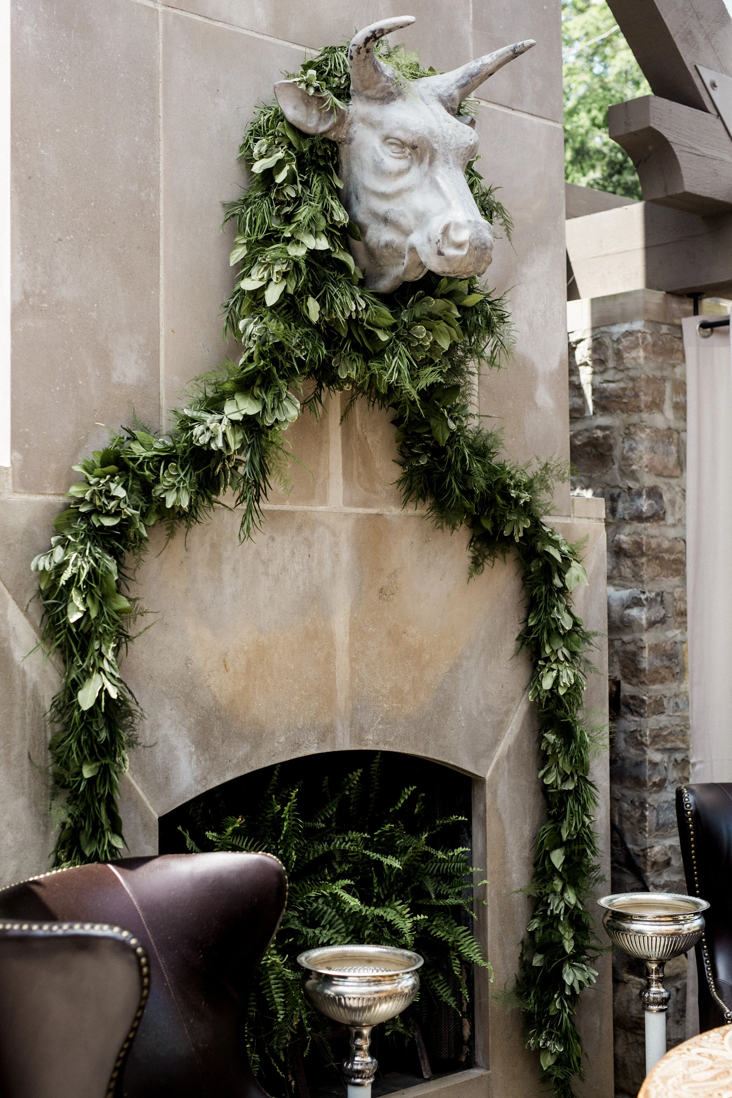Bull head sculpture above a stone fireplace with a decorative green garland draped around it, two leather chairs, and metal vessels near the fireplace.