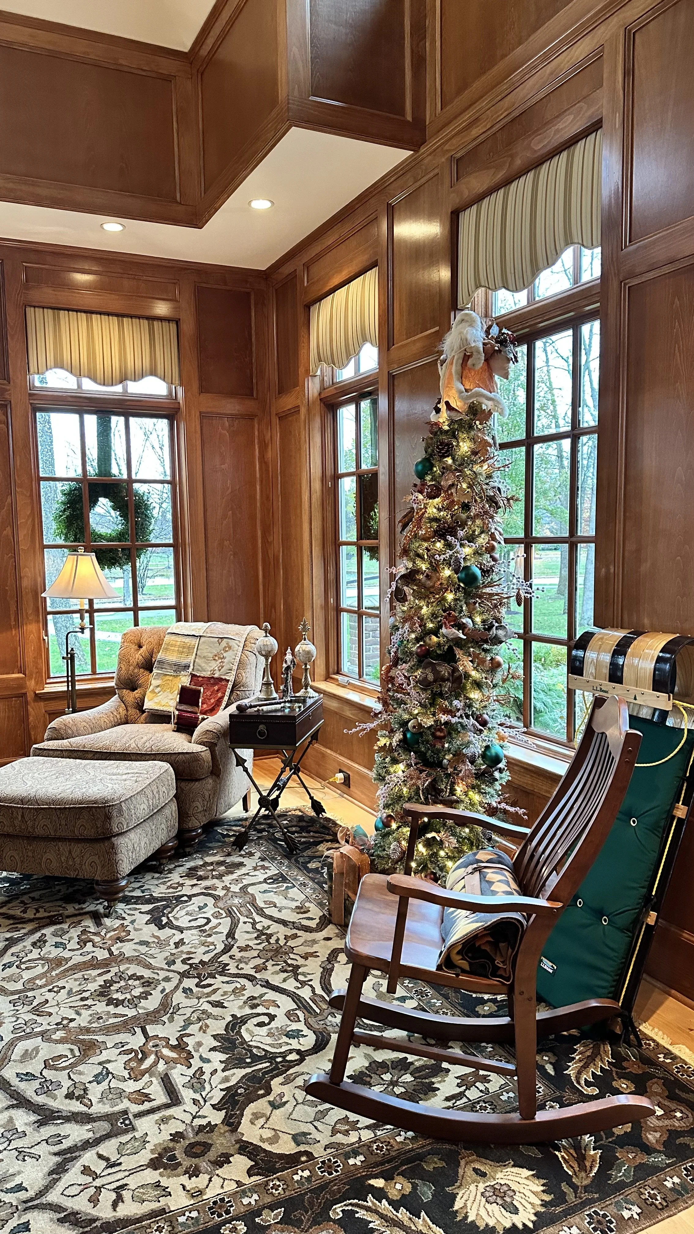 A cozy living room corner decorated for Christmas with a tall, artificial Christmas tree adorned with ornaments and a Santa hat on top, a rocking chair with a quilt draped over it, a beige armchair with an ottoman, a side table with decorations, a ta