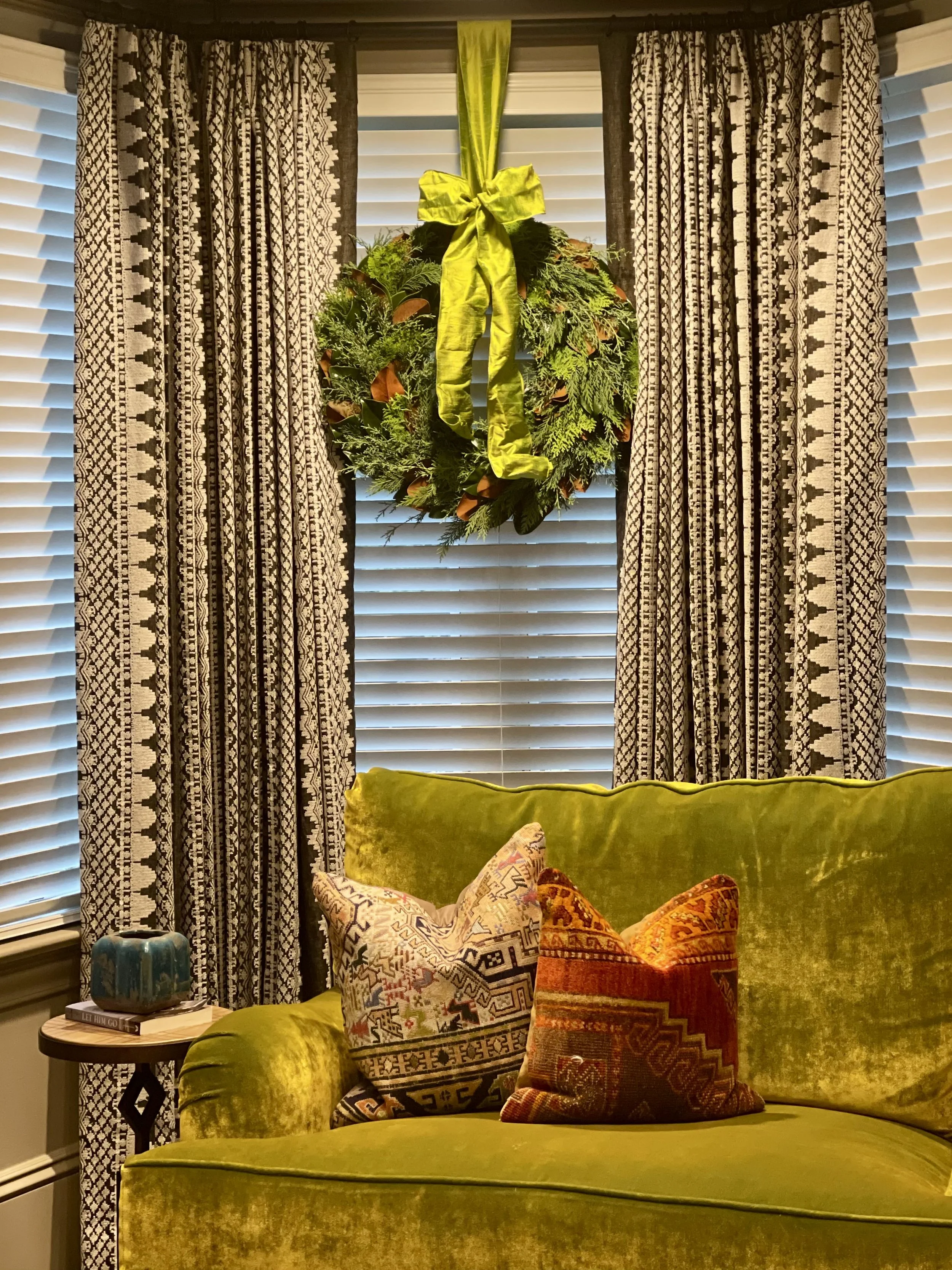 A living room scene with a green velvet sofa holding two decorative pillows with geometric and tribal patterns. Behind the sofa, there are large window blinds and patterned curtains. A hanging holiday wreath decorated with greenery and a yellow ribbo