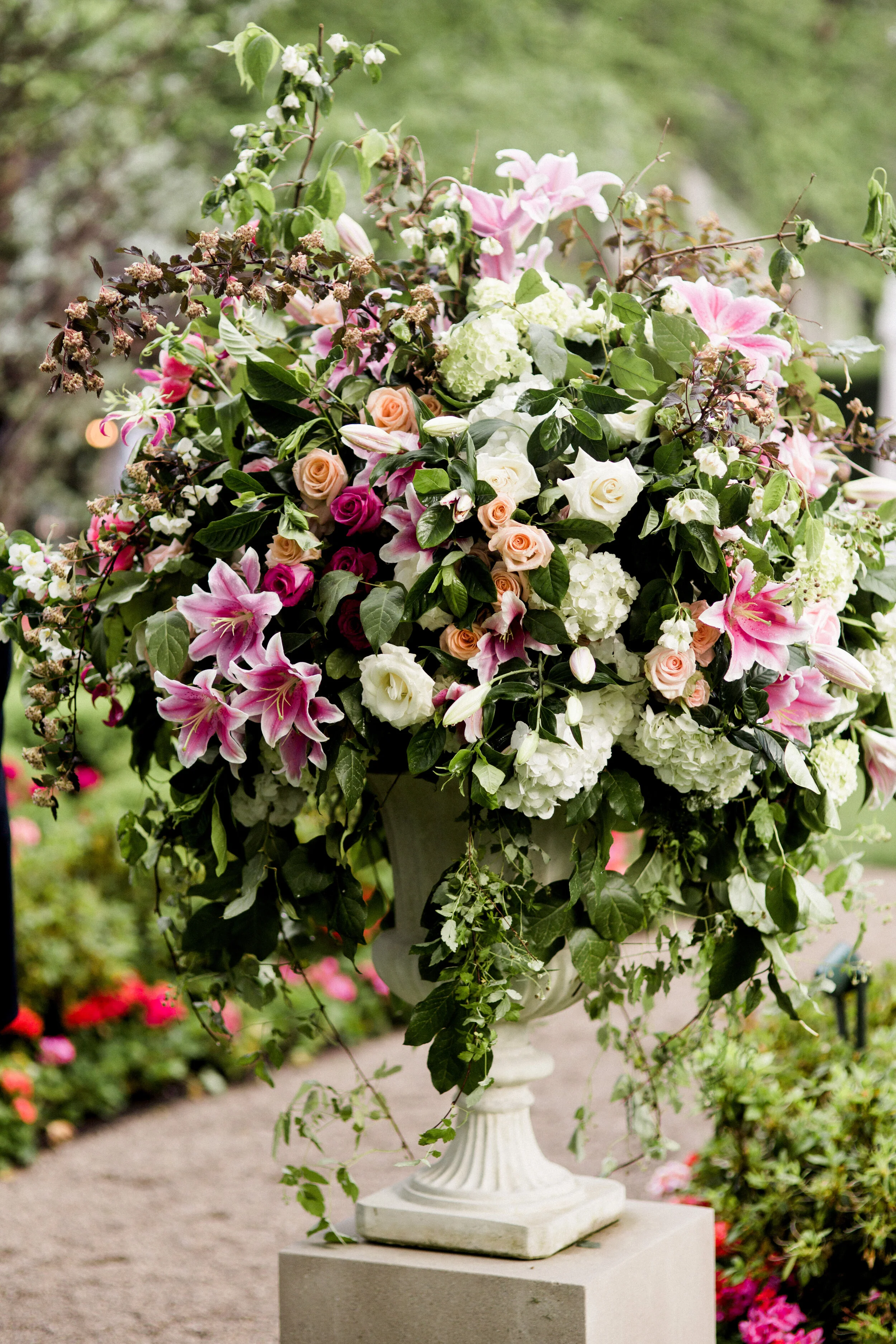 Large floral arrangement with pink lilies, roses, and greenery in an ornate planter outdoors.