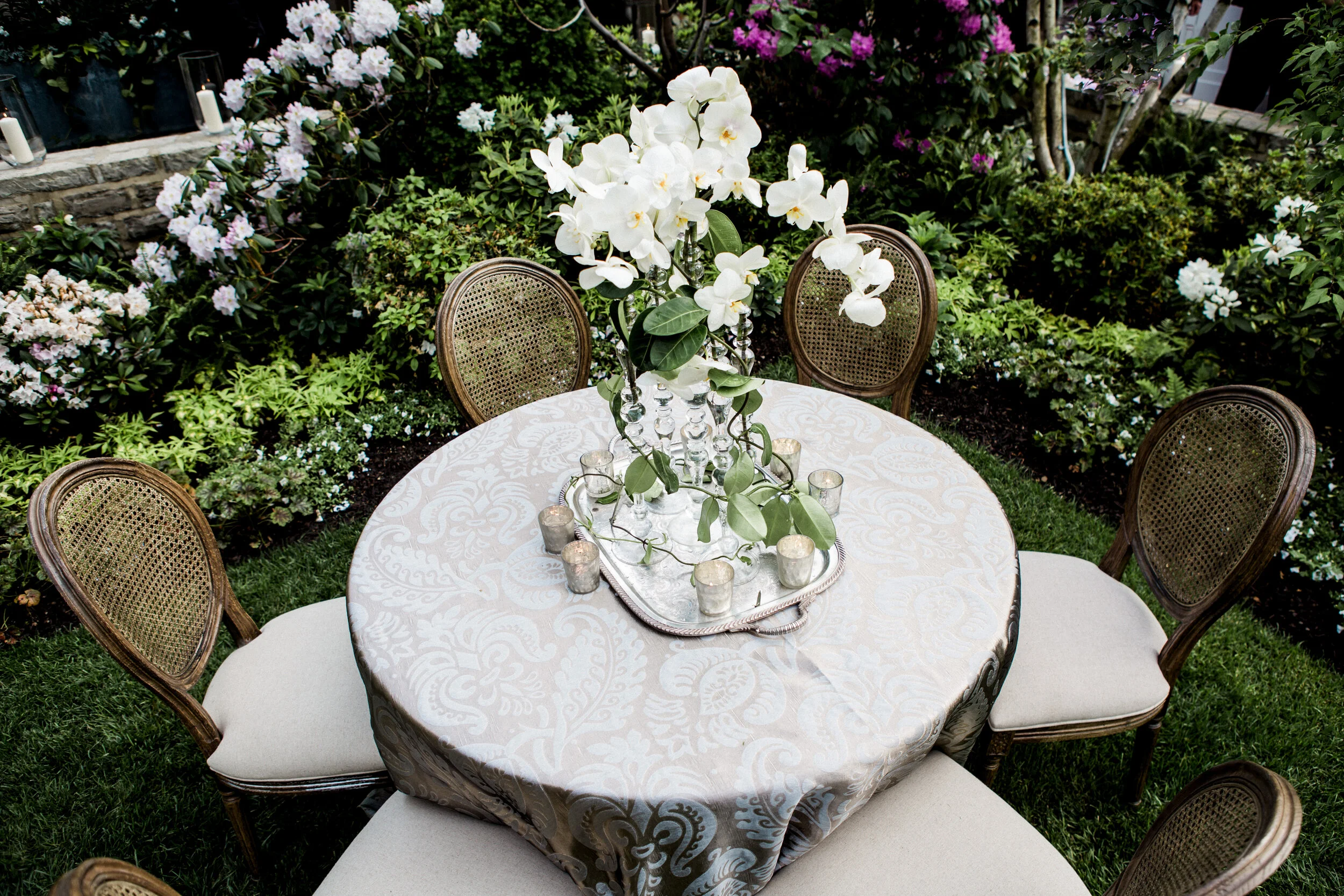 Elegant outdoor dining table with floral centerpiece and glass candle holders surrounded by vintage-style chairs, set in a lush garden backdrop.