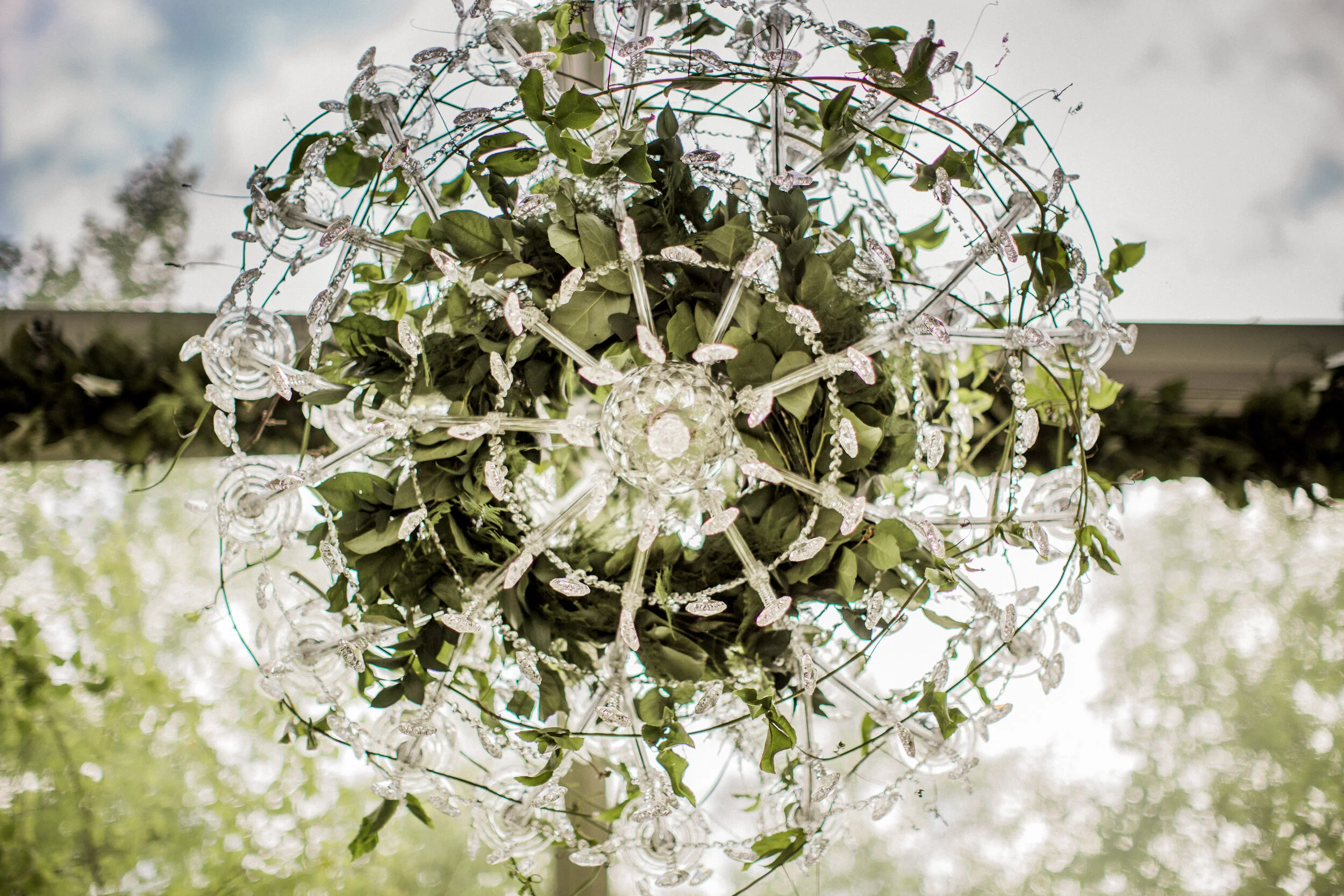 Elegant chandelier with greenery and crystal accents, set against a blurred outdoor background.