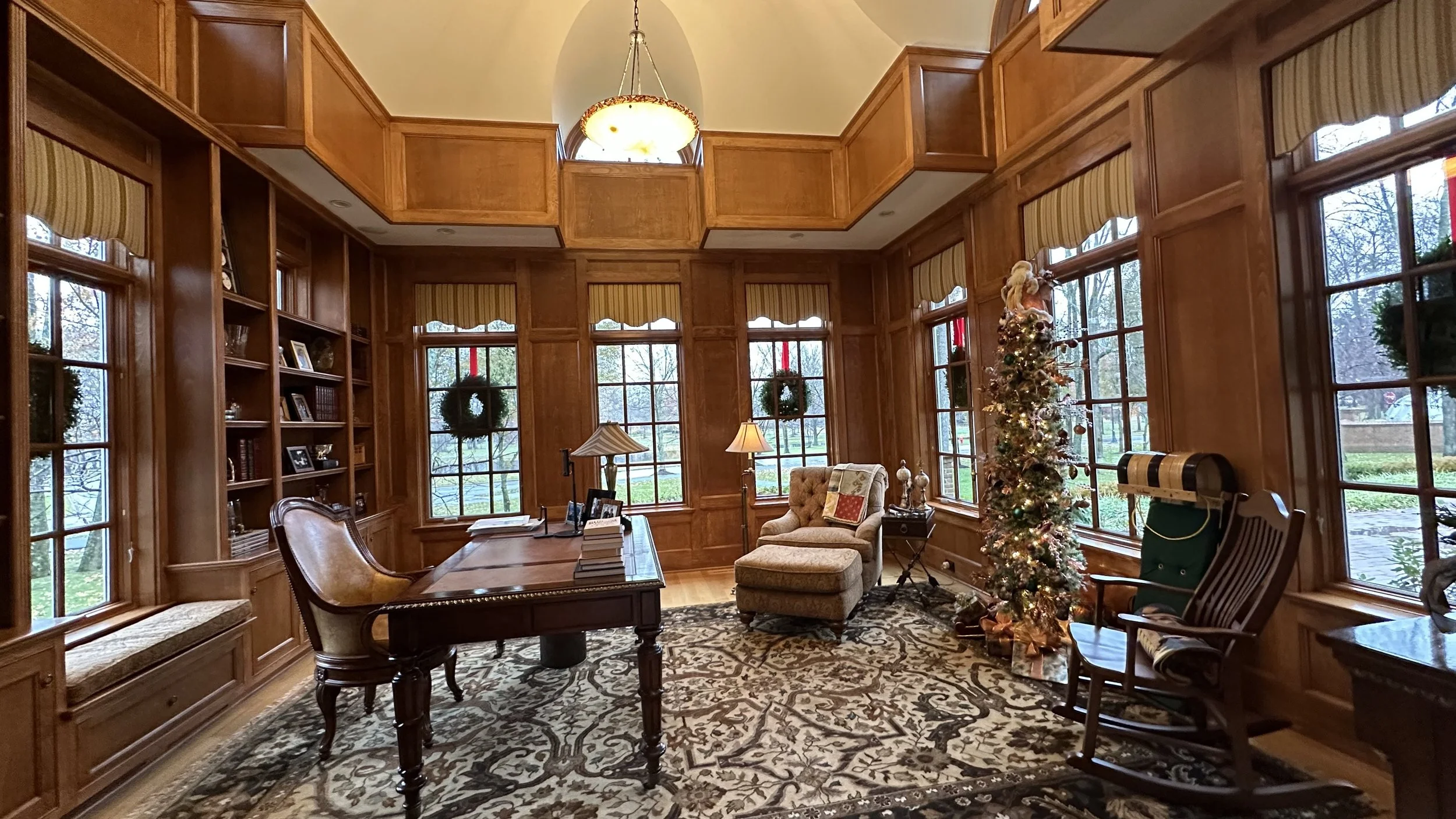 A cozy, wood-paneled home office decorated for Christmas with a decorated tree, wreaths on the windows, and holiday accents. The room has large windows, a desk with papers and a lamp, an armchair with a throw pillow, and a patterned area rug.